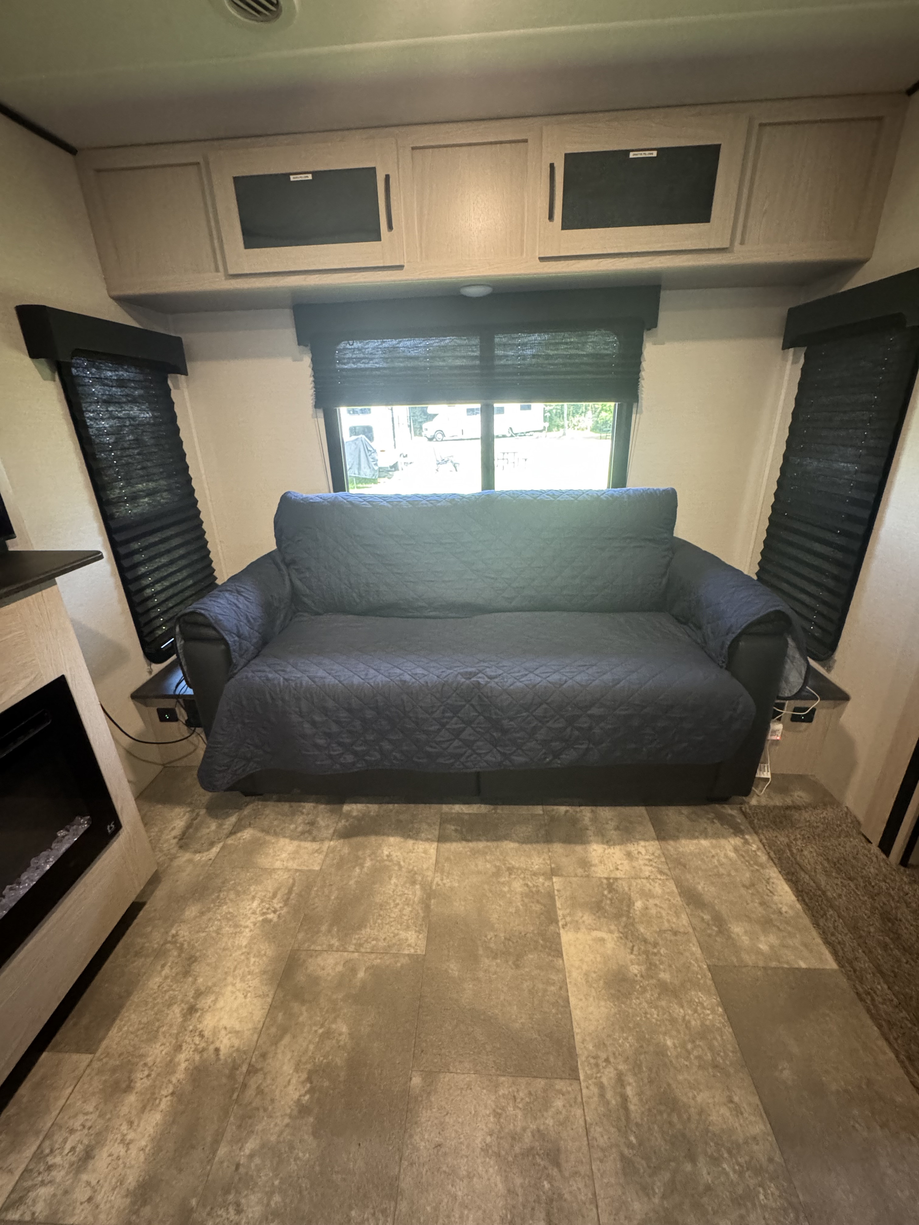 Spacious RV rental interior featuring a dark blue quilted sofa, overhead cabinets, and large windows with blinds.. Coachmen Apex 2022