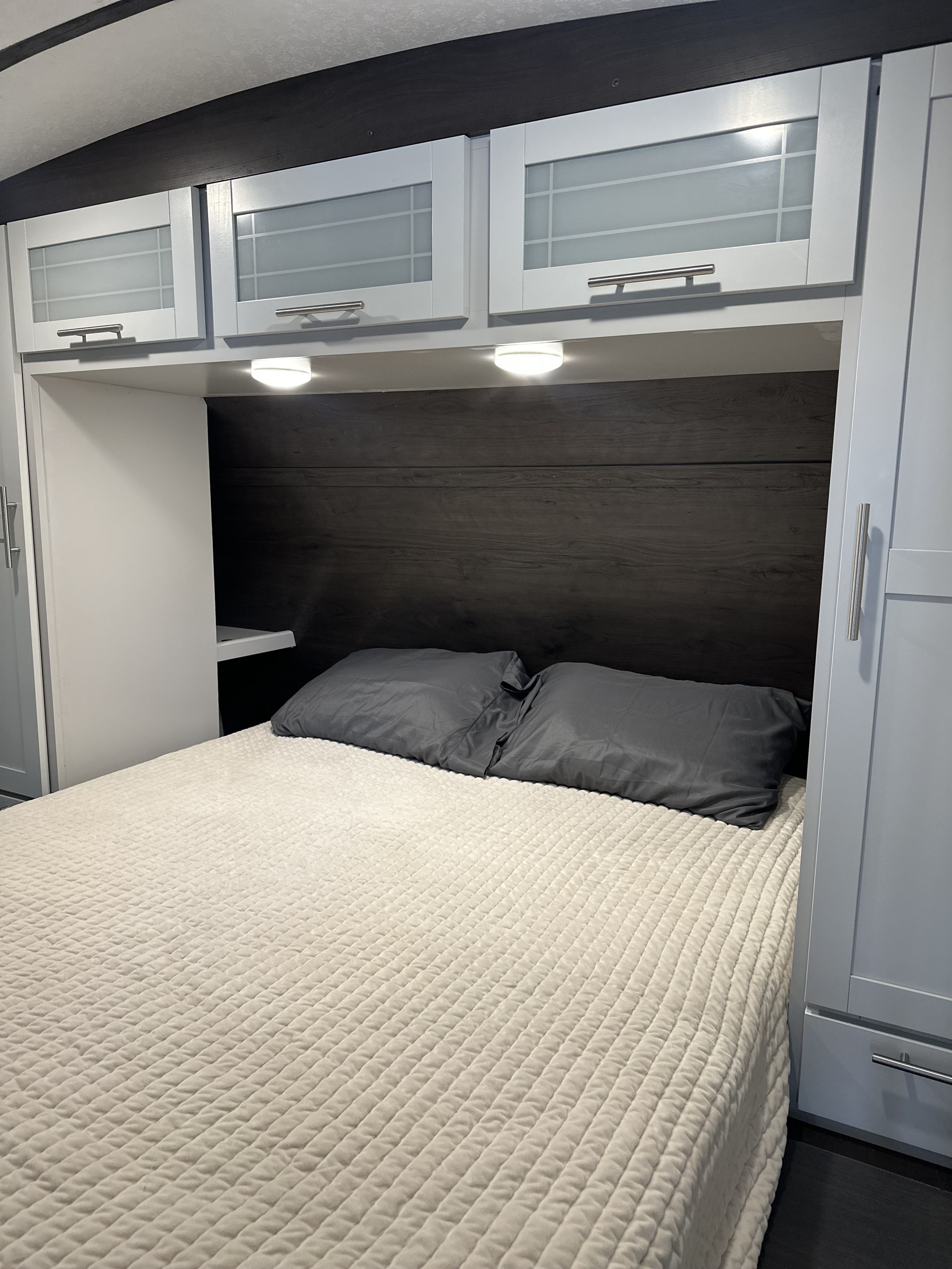 A cozy RV rental bedroom features a quilted bed, dark wood headboard, light grey cabinets, & built-in lights.. Keystone RV Bullet Ultra Lite 2021