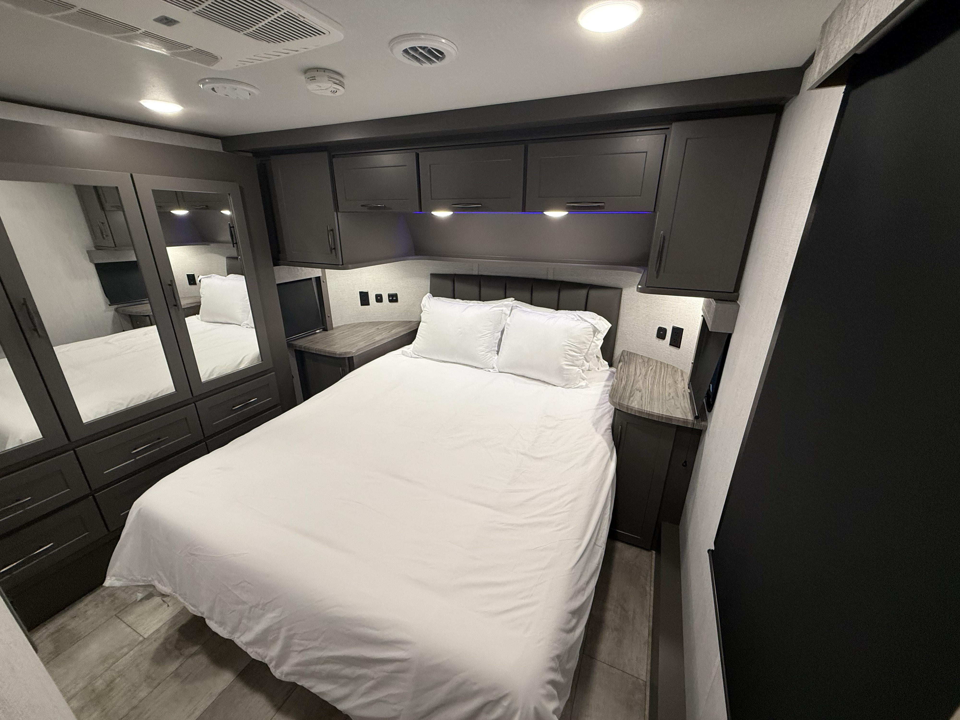 Spacious, modern RV rental bedroom with white sheets, dark wood cabinets, and bright overhead lighting, plus mirrored wardrobe doors.. Grand Design Grand Design Fifth Wheel 2024
