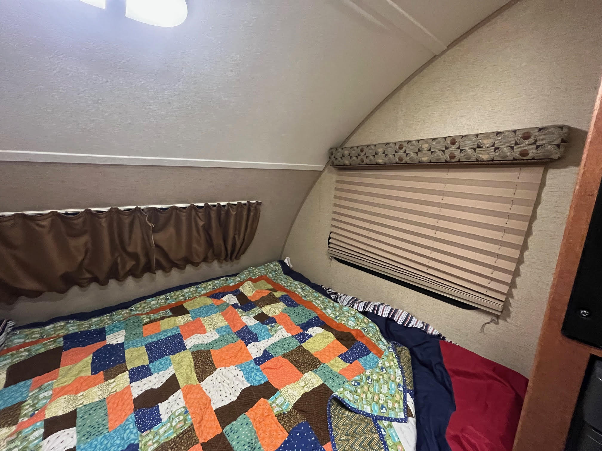 Interior of a comfortable RV rental bedroom, featuring a vibrant patchwork quilt, window blinds, and bright ceiling light.. R-Pod R-Pod Trailer 2015