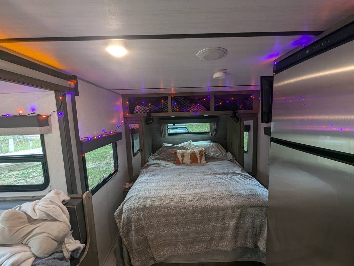 A comfy RV rental bedroom, featuring a large bed, decorative purple & orange string lights, and a stainless steel fridge.. Forest River Surveyor Legend 19RBLE 2022