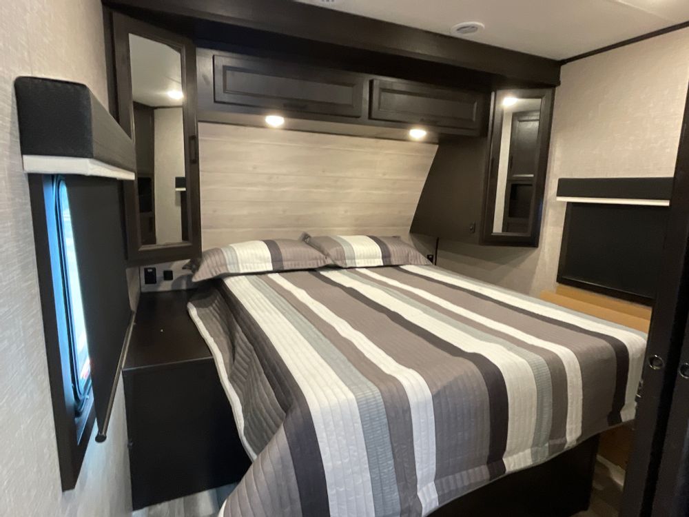 Comfortable RV rental bedroom featuring a queen bed with striped bedding, dark cabinets, and windows. A cozy private space for travel.. Jayco Eagle HT 2023