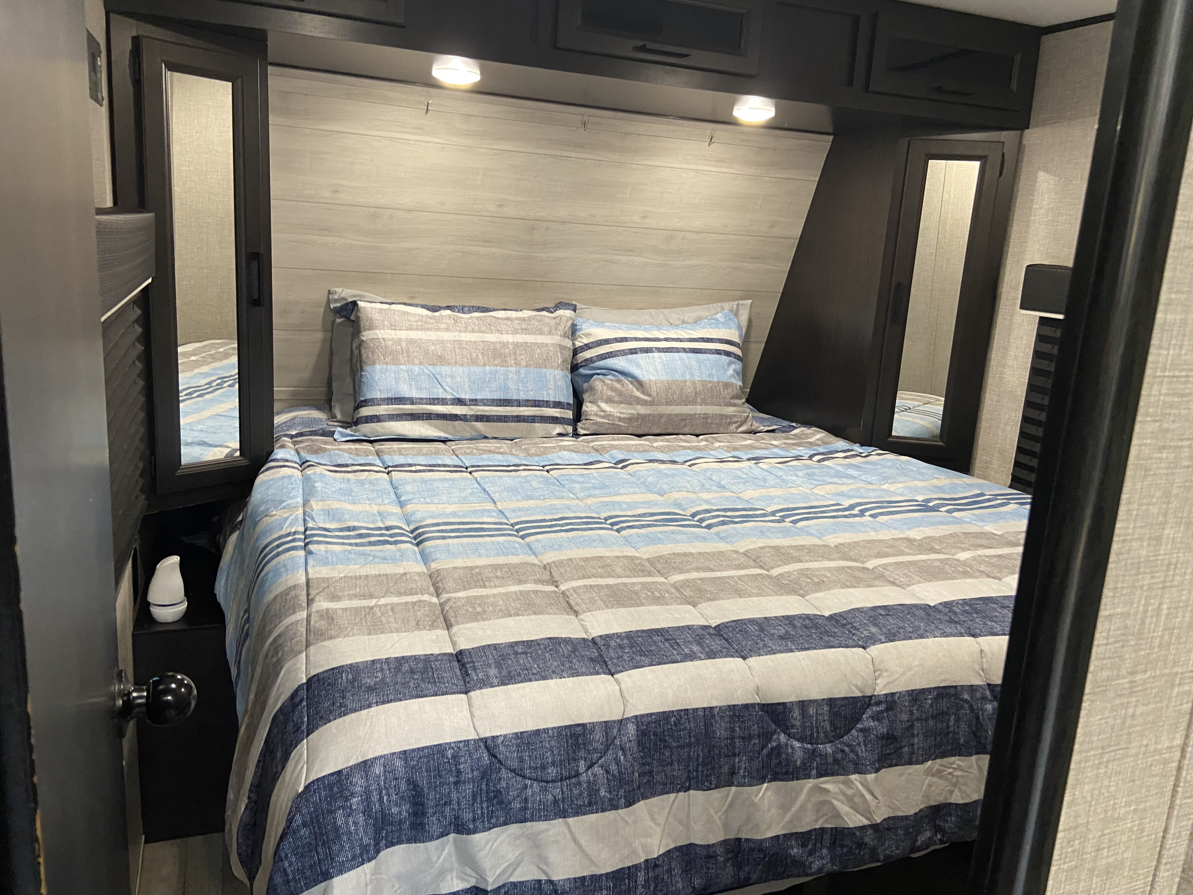 Cozy RV rental bedroom interior, featuring blue, gray, and white striped bedding, overhead lights, and mirror storage.. Jayco jayflight 2022