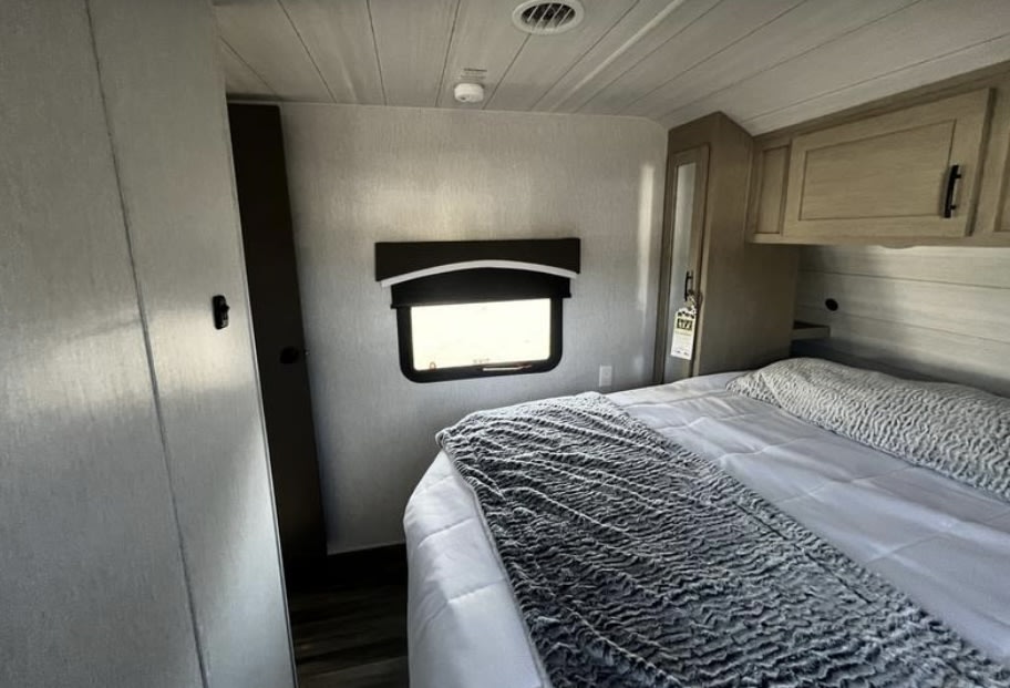 Cozy RV rental bedroom, featuring a large bed with gray and white bedding, window, and built-in storage cabinets. Ideal for travel.. Forest River Eco 2024