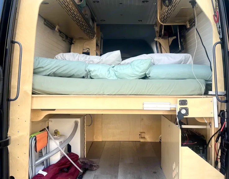 Functional RV rental interior: raised bed with bedding, ample storage, water tank, and electricals visible below.. Mercedez Sprinter 2014