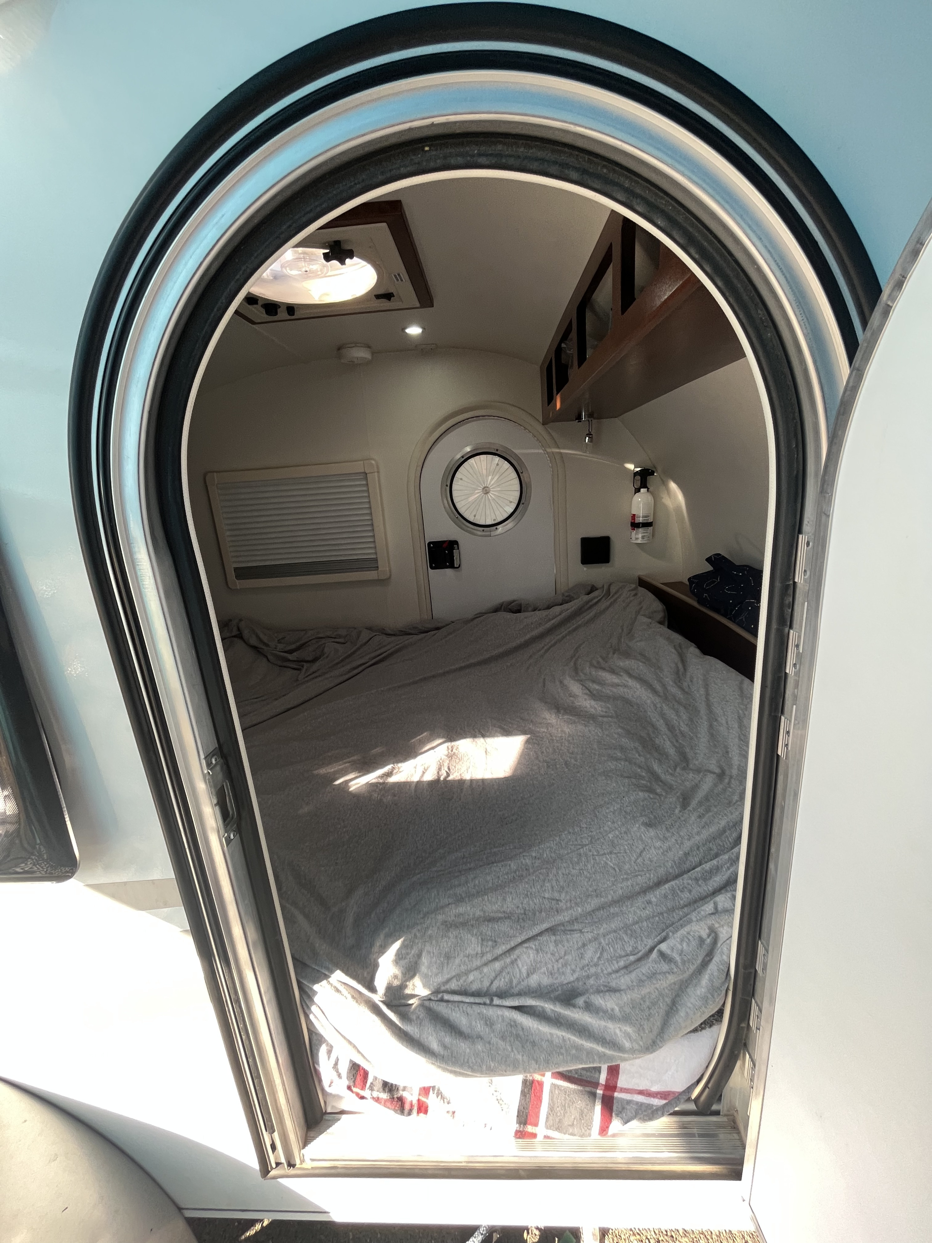 Step into a spacious RV rental bed area, seen through an arched door. Features include a comfy grey bed, small round door, and overhead storage.. T@G T@G Teardrop Trailer 2015