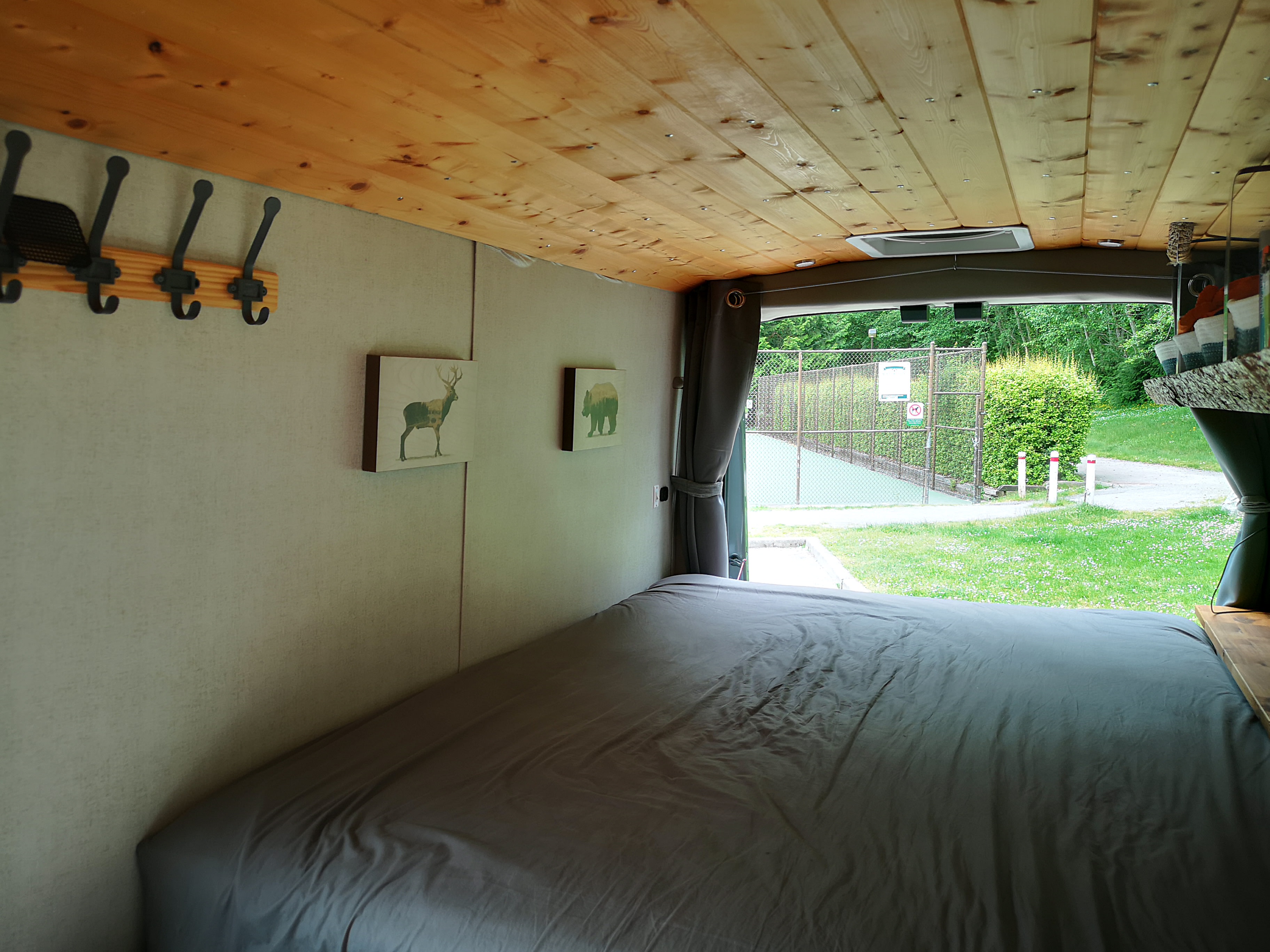 Cozy RV rental interior shows a comfortable bed, wooden ceiling, and a green outdoor view from the open back.. Ford Transit 2022