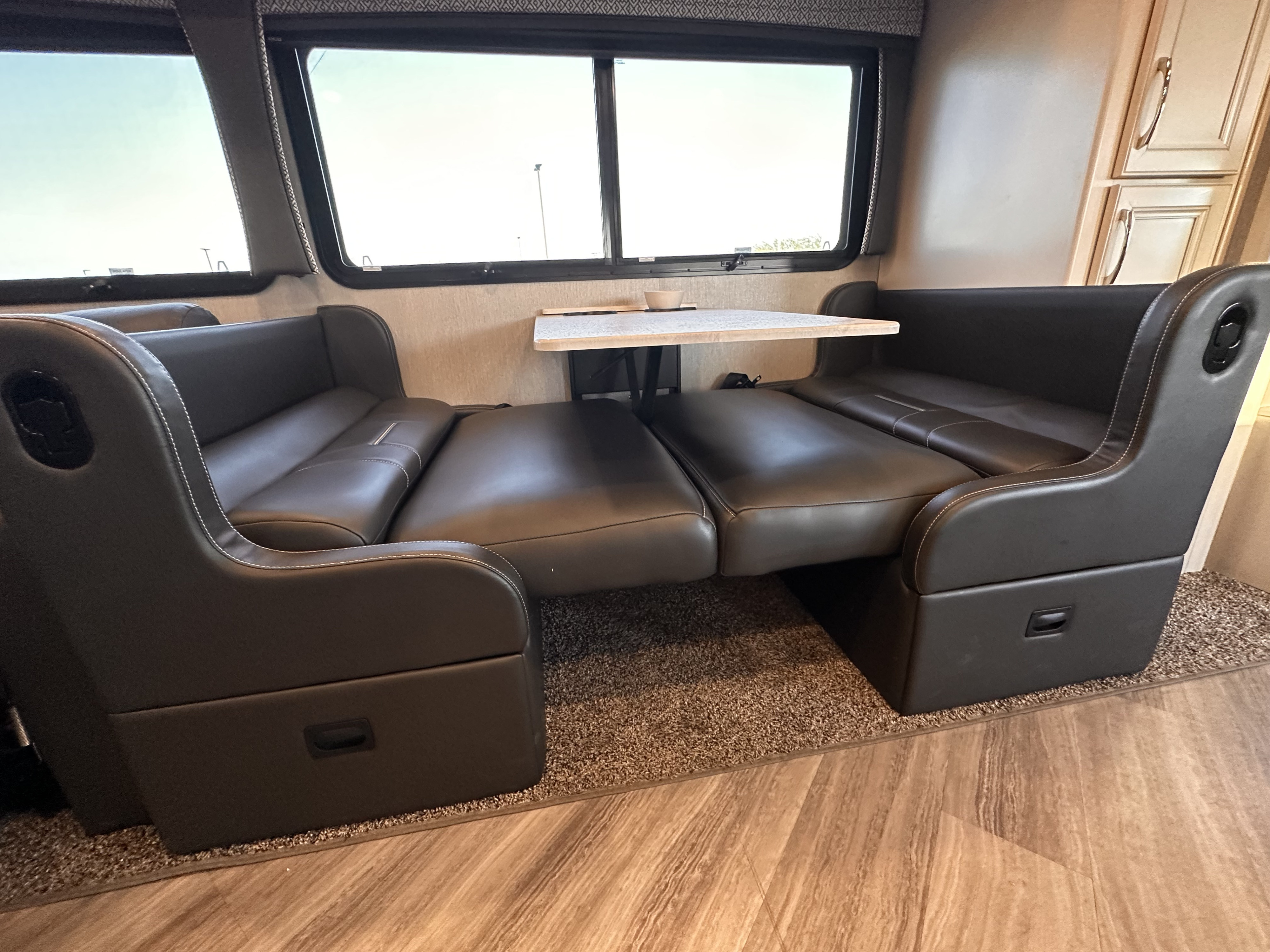 Inside an RV rental: dark upholstered dinette with a table, convertible to a bed. Large windows overlook the scenery.. Holiday Rambler Vacationer 2022