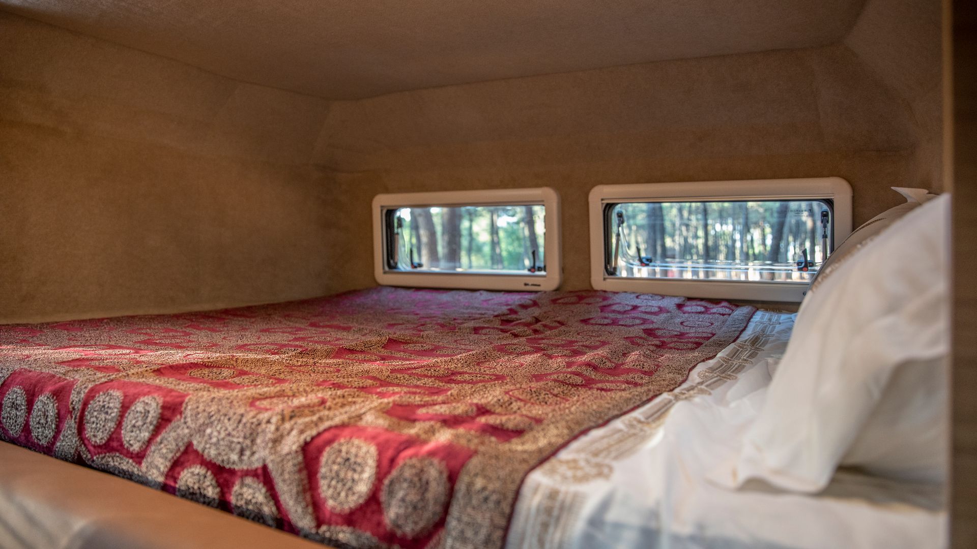 RV rental interior: comfortable bed with a patterned blanket, offering serene forest views from its windows.. F-150 Lightning / Hotomobil Gladiator XL with roof tent 2023