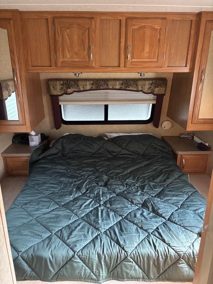 Cozy RV rental bedroom featuring a large bed with a green comforter, wooden overhead cabinets, and a window. Perfect for your travels.. Mckenzie Starwood LX 2008