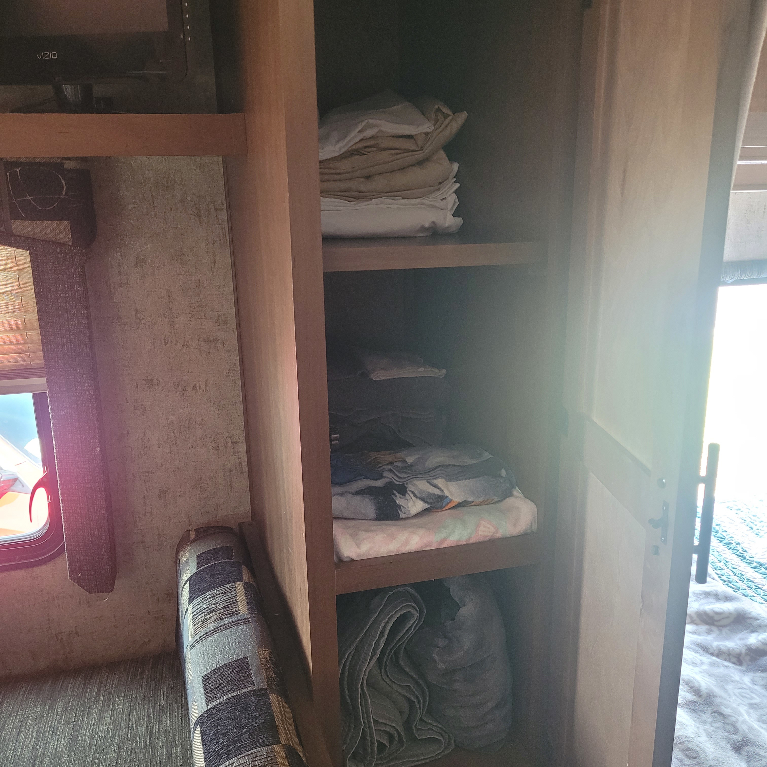 Inside an RV rental, a compact storage cabinet holds folded linens, with a TV and window visible, showcasing convenient storage.. Outdoors Rv Manufacturing Black Rock 2017
