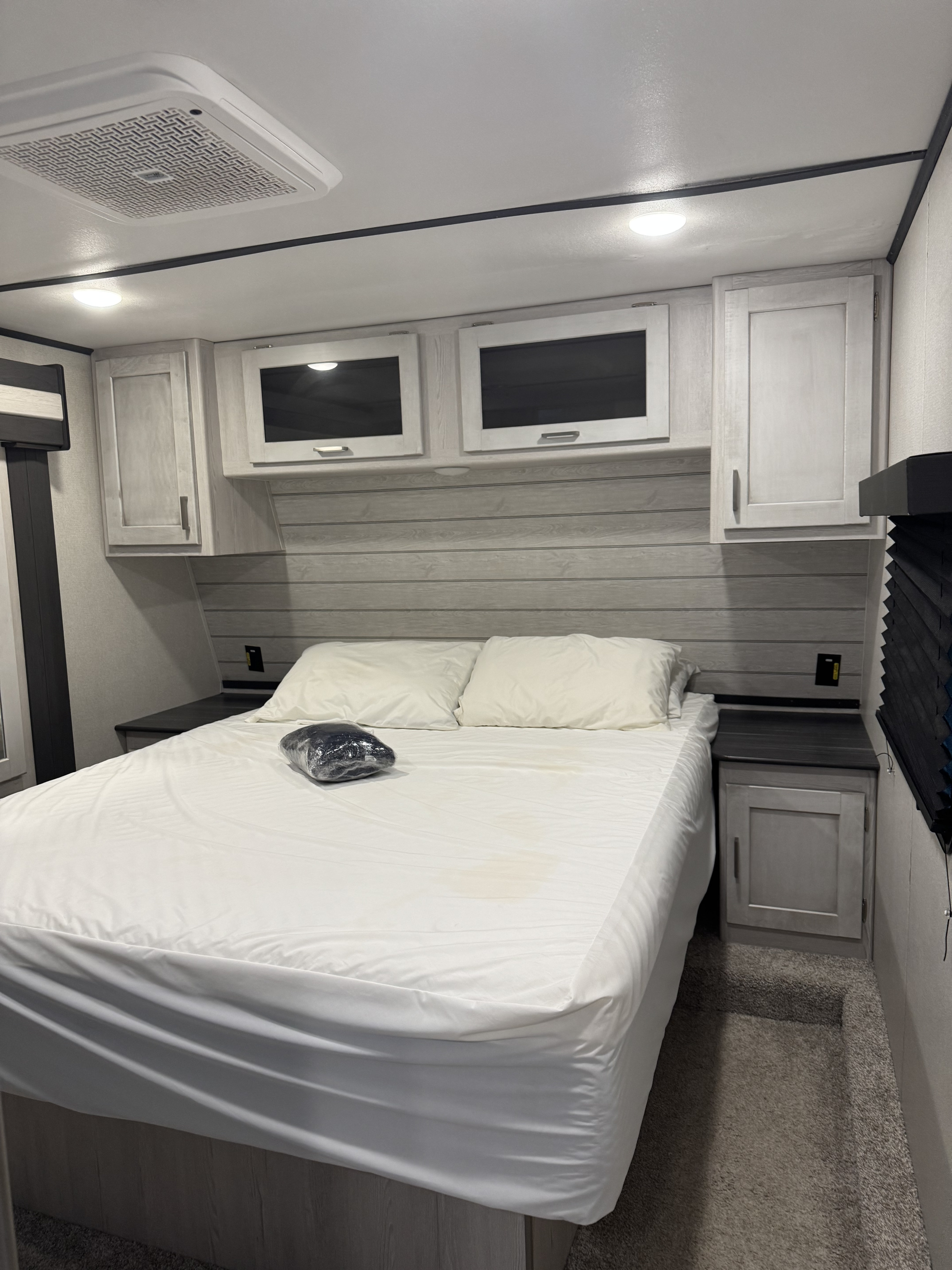 This RV rental bedroom features a white queen bed with new bedding, overhead cabinets, and a ceiling AC unit in a light wood-paneled space.. Keystone Sprinter 2022