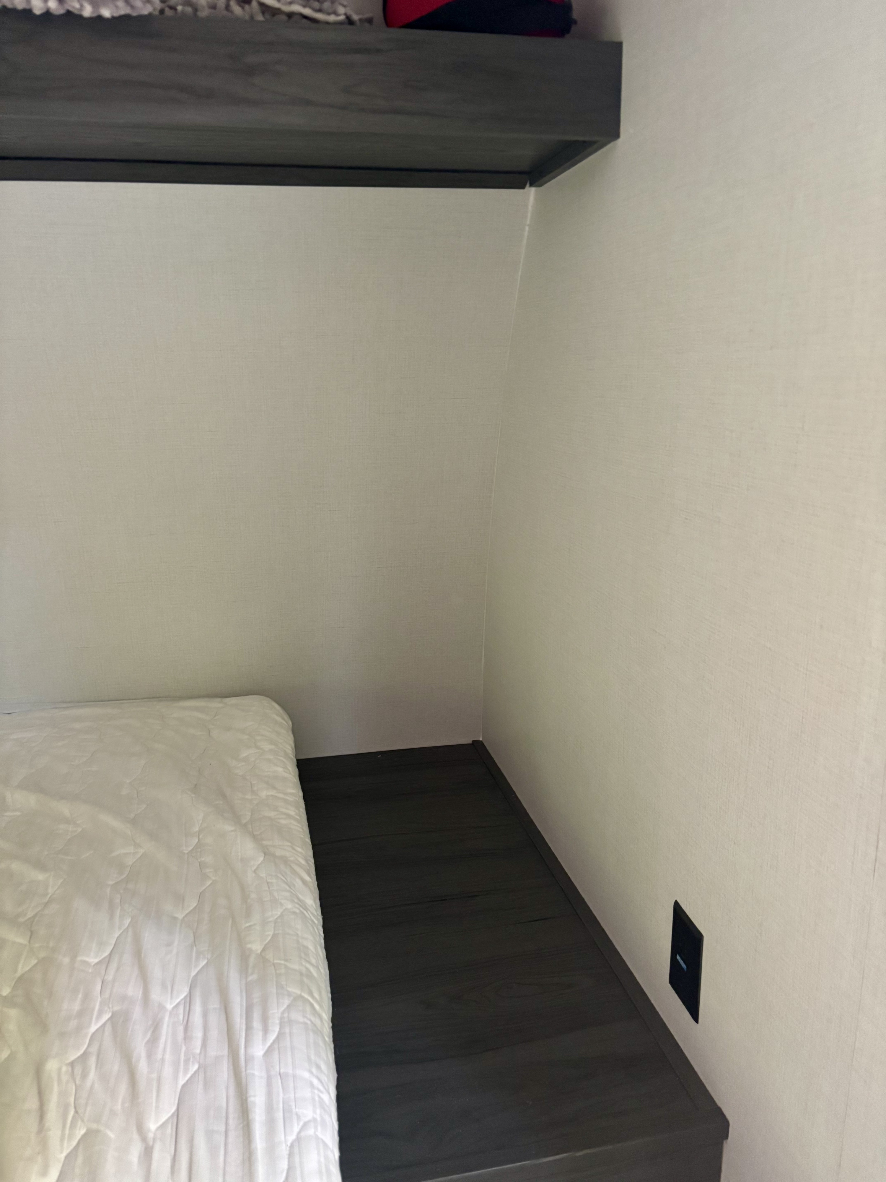 Inside an RV rental: a compact bedroom with a white mattress, overhead shelf, and wall charging port.. Dutchman Aspen Trail 2025