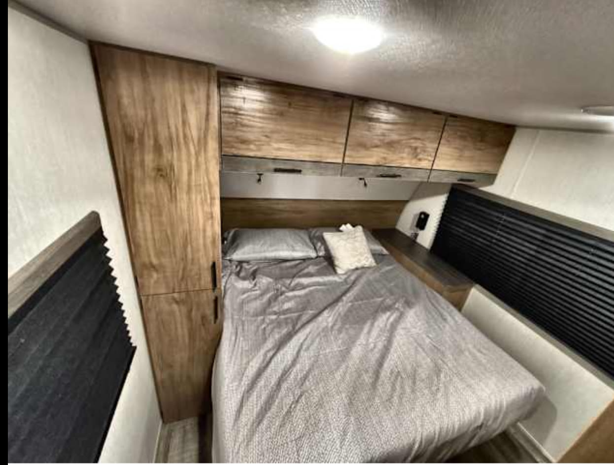 RV rental bedroom: A comfortable bed with grey bedding, wood-tone cabinets, and window blinds.. Forest River Campsite Reserve 26CJ 2025