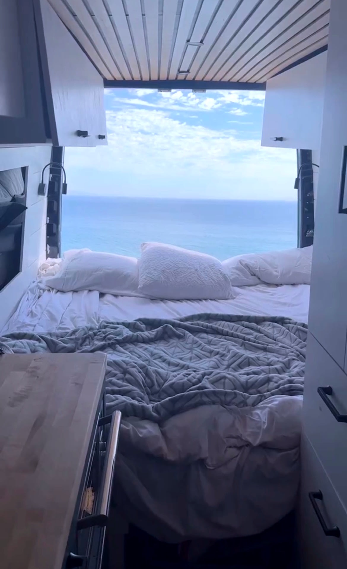 Inside an RV rental, a cozy bed faces open doors revealing a panoramic blue ocean and sky view. Perfect for scenic travel!. Mercedes-Benz Sprinter 2017