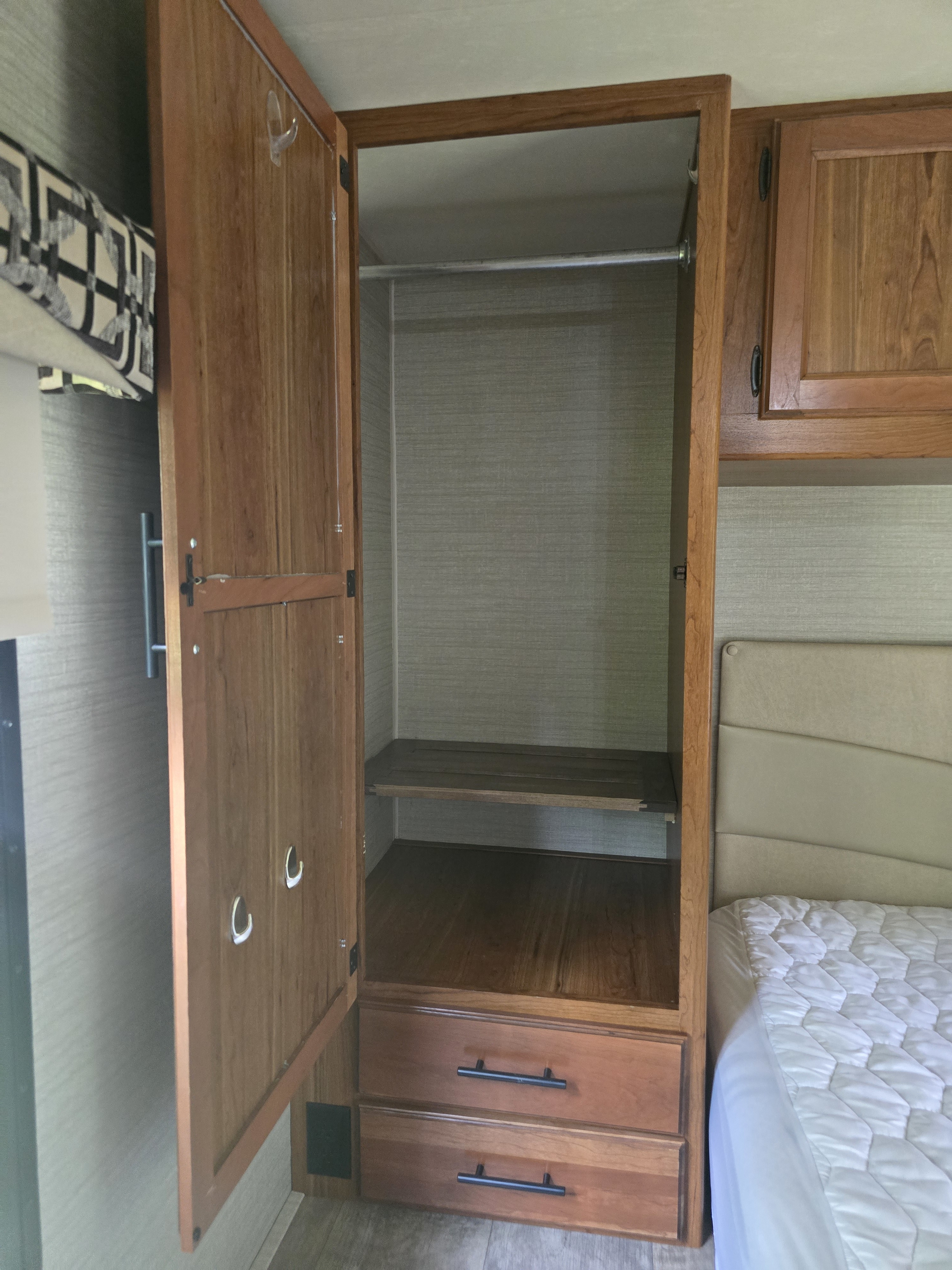 RV rental bedroom features a compact wooden wardrobe with a hanging rod, shelf, and drawers, beside a cozy bed.. Jayco Redhawk 2017