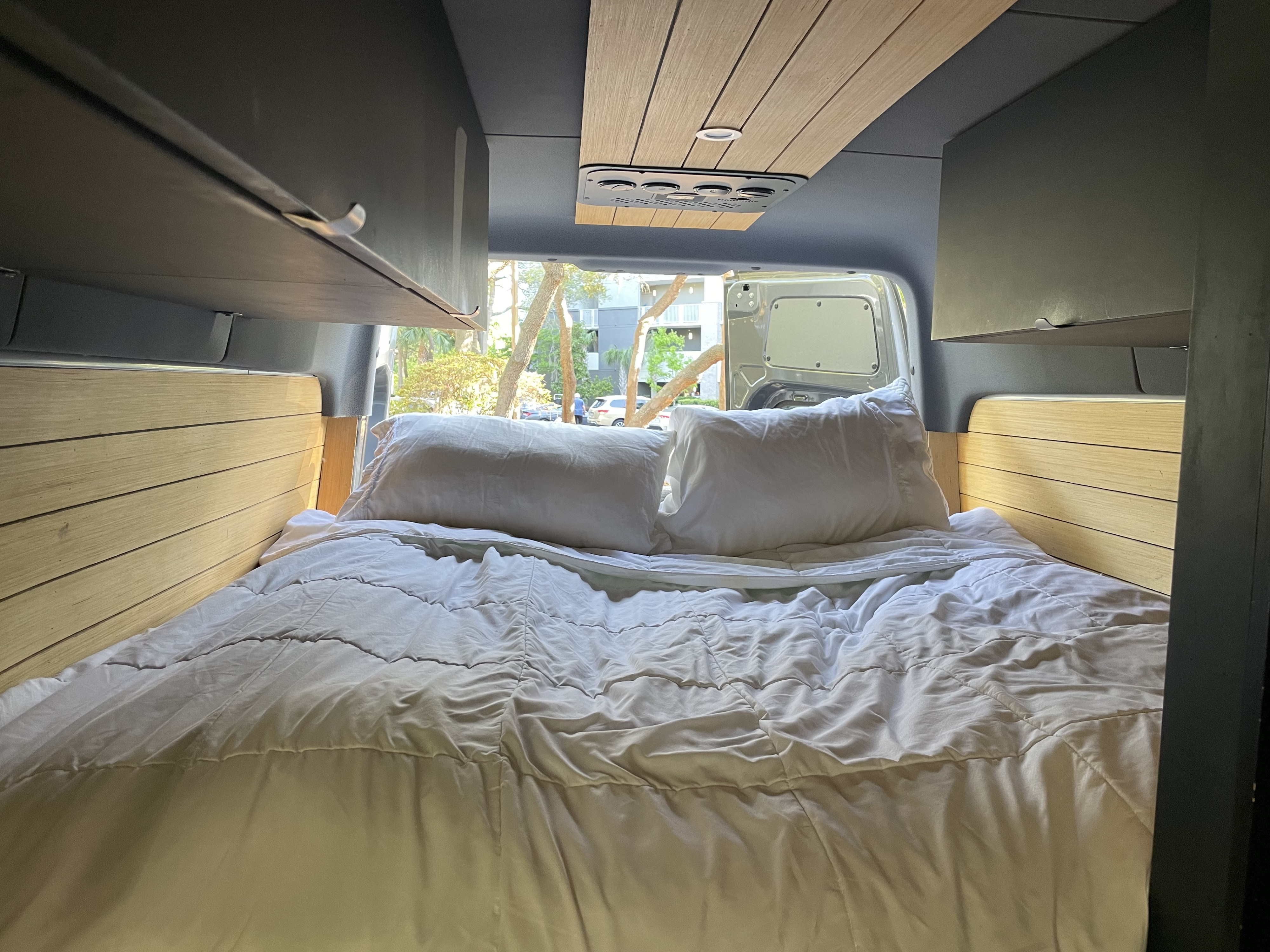 Comfortable RV rental interior with a cozy bed, white bedding, wooden accents, and open rear doors revealing a scenic view.. Mercedes Benz Sprinter 2500 4x4 Off Grid Conversion 2020