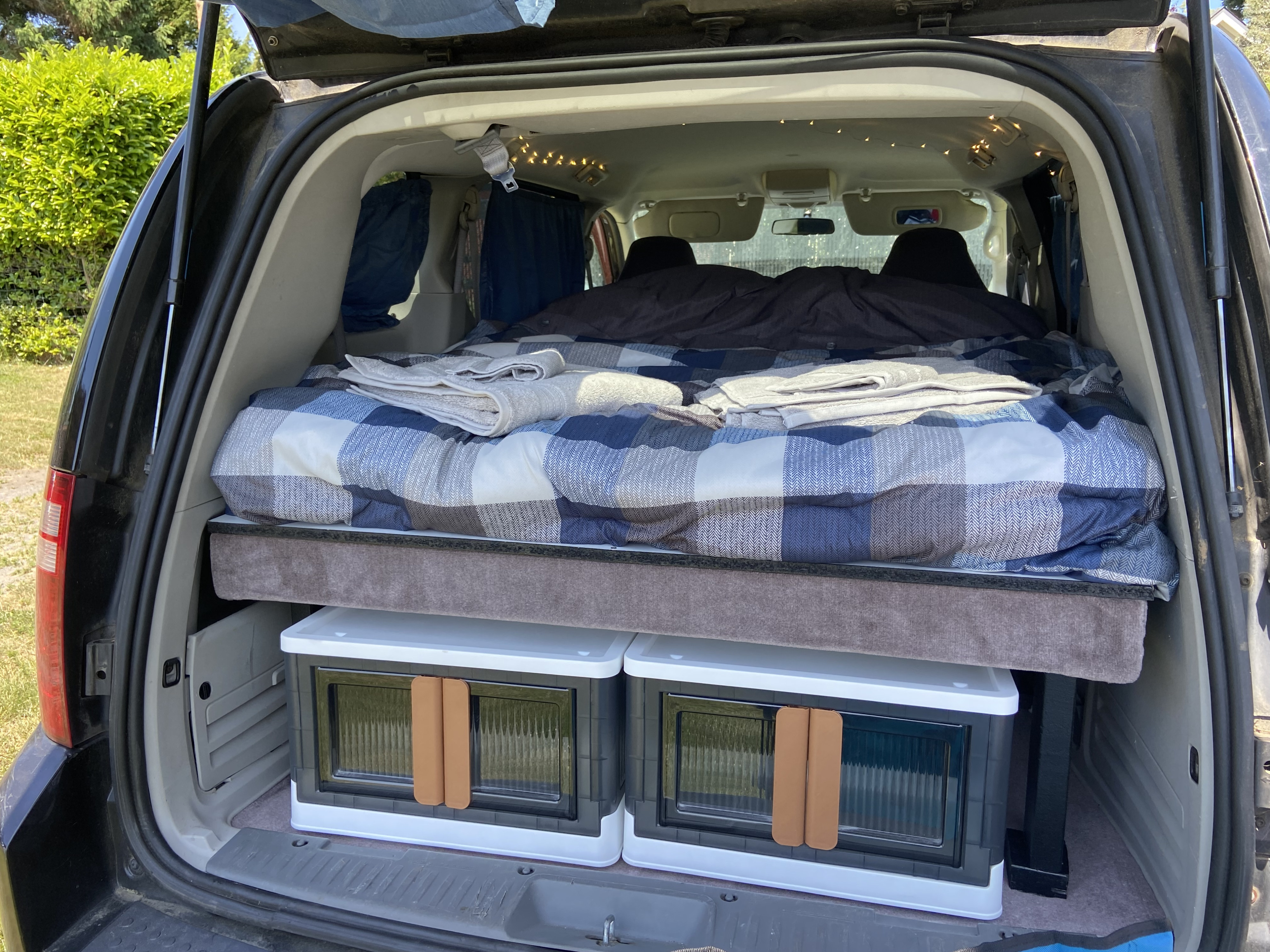 Cozy RV rental setup: inside view of a camper van with bed, storage bins, and string lights for camping.. Dodge Grand Caravan 2008