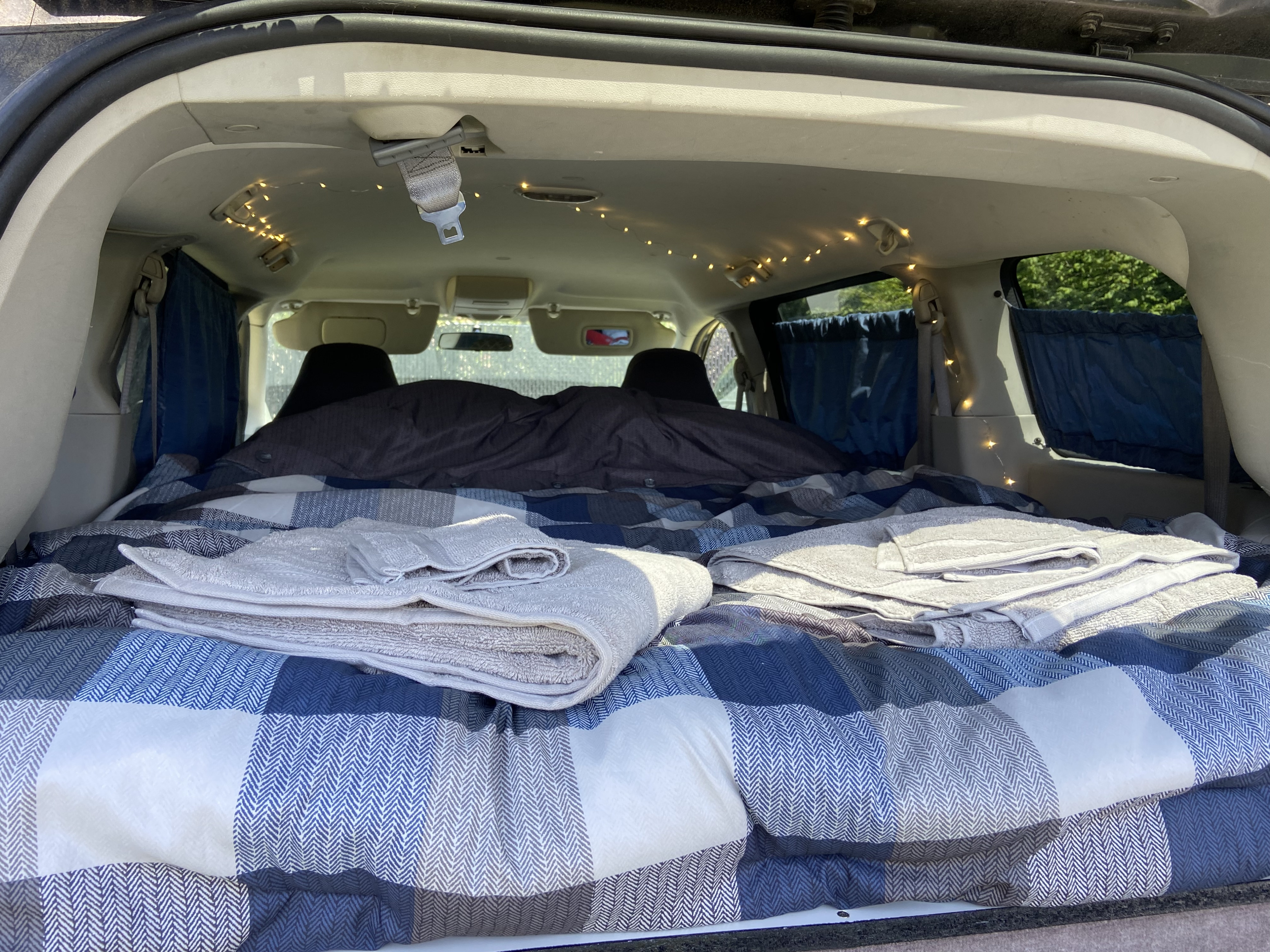 Inside an RV rental setup: a cozy bed with plaid blankets, towels, and string lights create a comfy car camping retreat.. Dodge Grand Caravan 2008