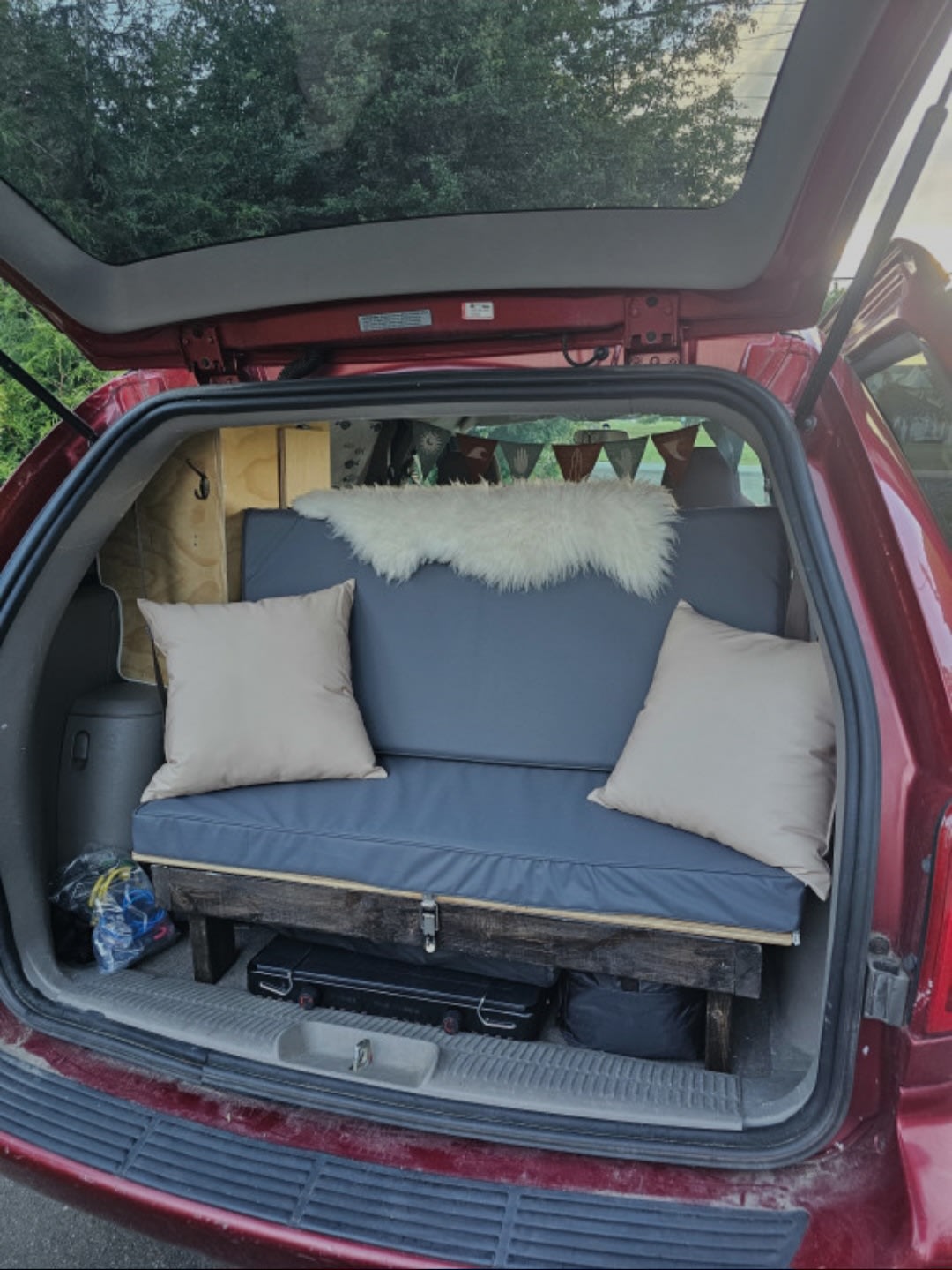 Red minivan's tailgate open, revealing a DIY camper conversion with a blue cushioned bench bed. Ideal for RV rental travel.. Dodge Caravan 2006