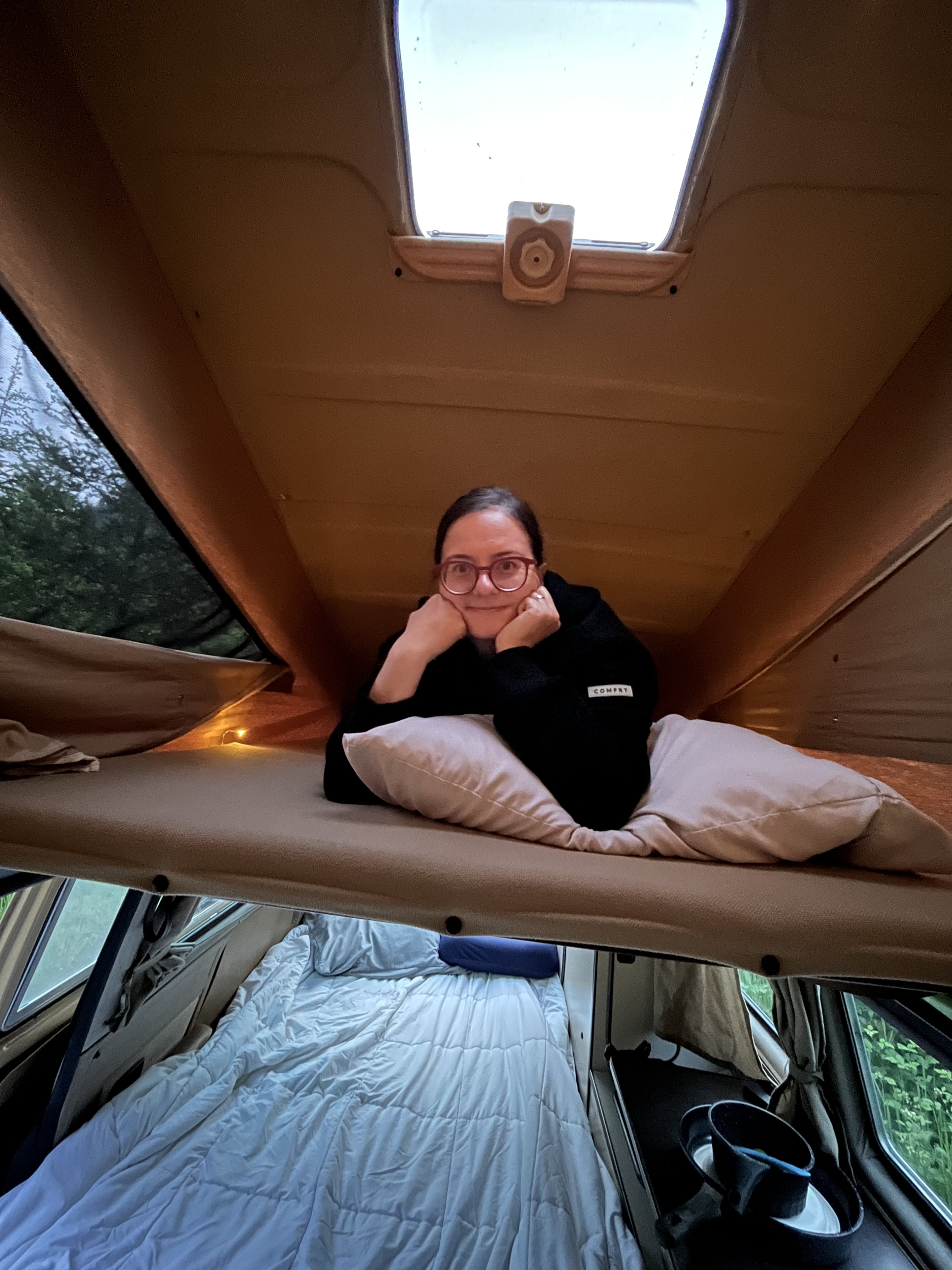 Enjoying a comfortable **RV rental**: A woman smiles from the pop-top bed, with a lower bed and outdoor view.. Volkswagen Westfalia 1985