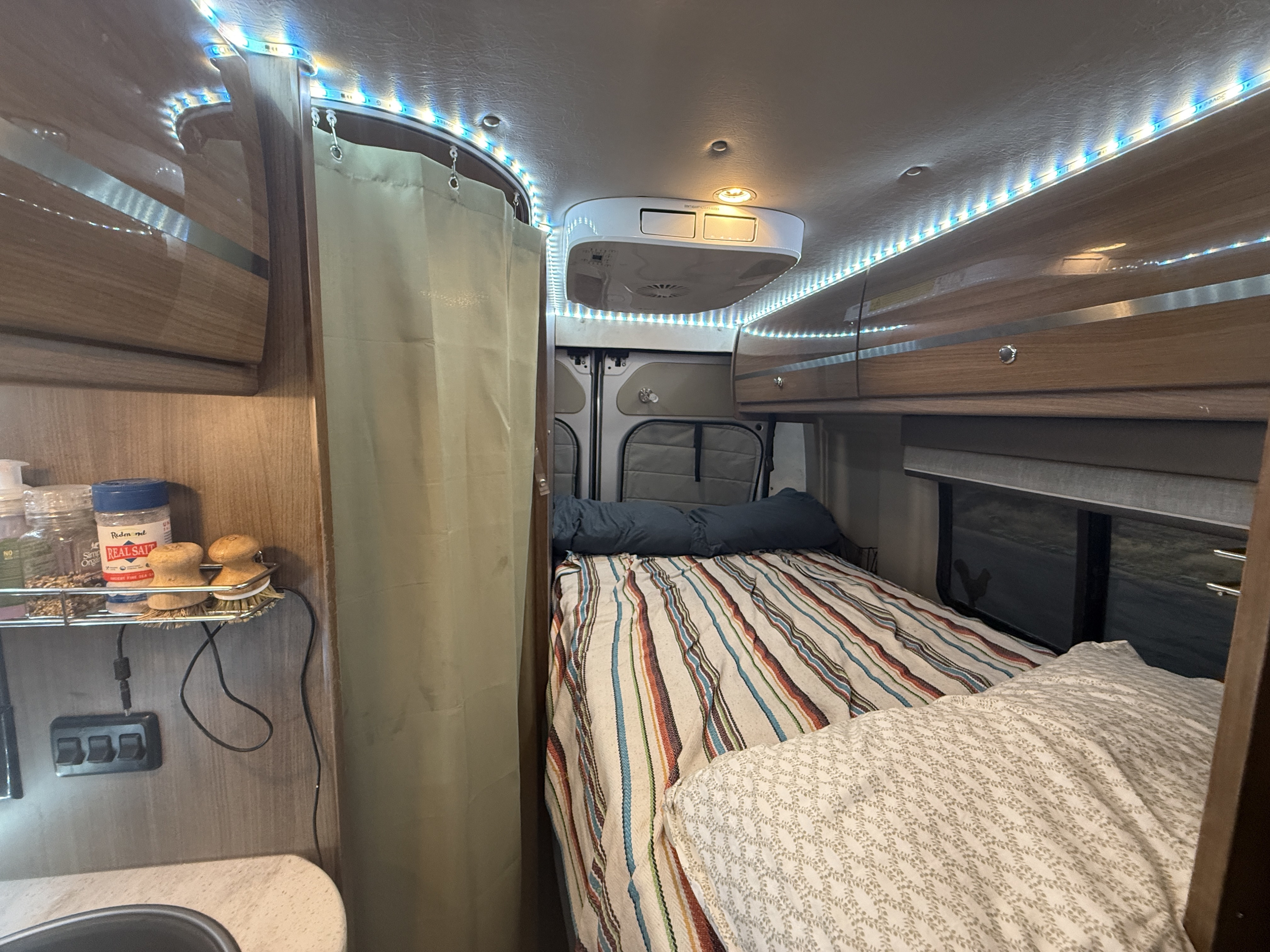 Cozy RV rental interior with bed, overhead cabinets, LED lighting, and a compact kitchen area.. Winnebago Travato 2016