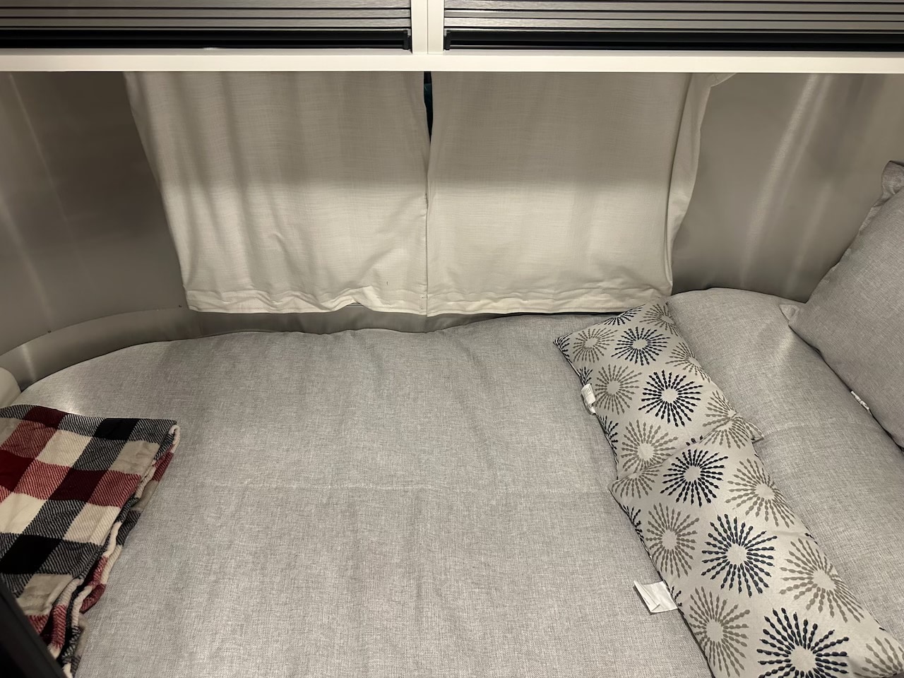 Cozy RV rental bed features light gray linens, a red plaid blanket, and patterned pillows. Perfect for a comfortable travel trailer trip.. Airstream Bambi 2022