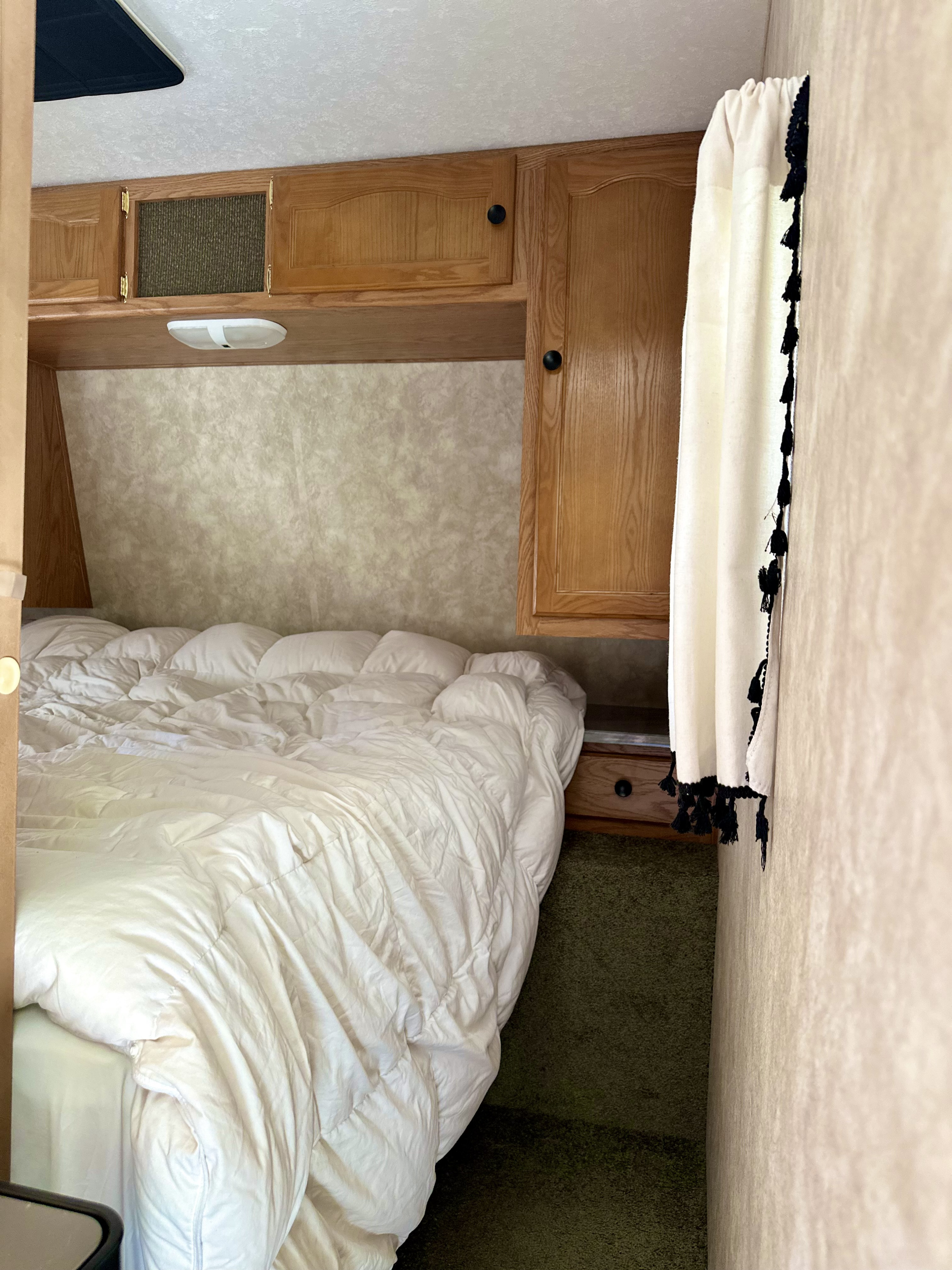 Cozy RV rental bedroom with wooden cabinets, light fixture, white bedding, green carpet, and a tassel curtain.. Keystone RV Springdale 2007