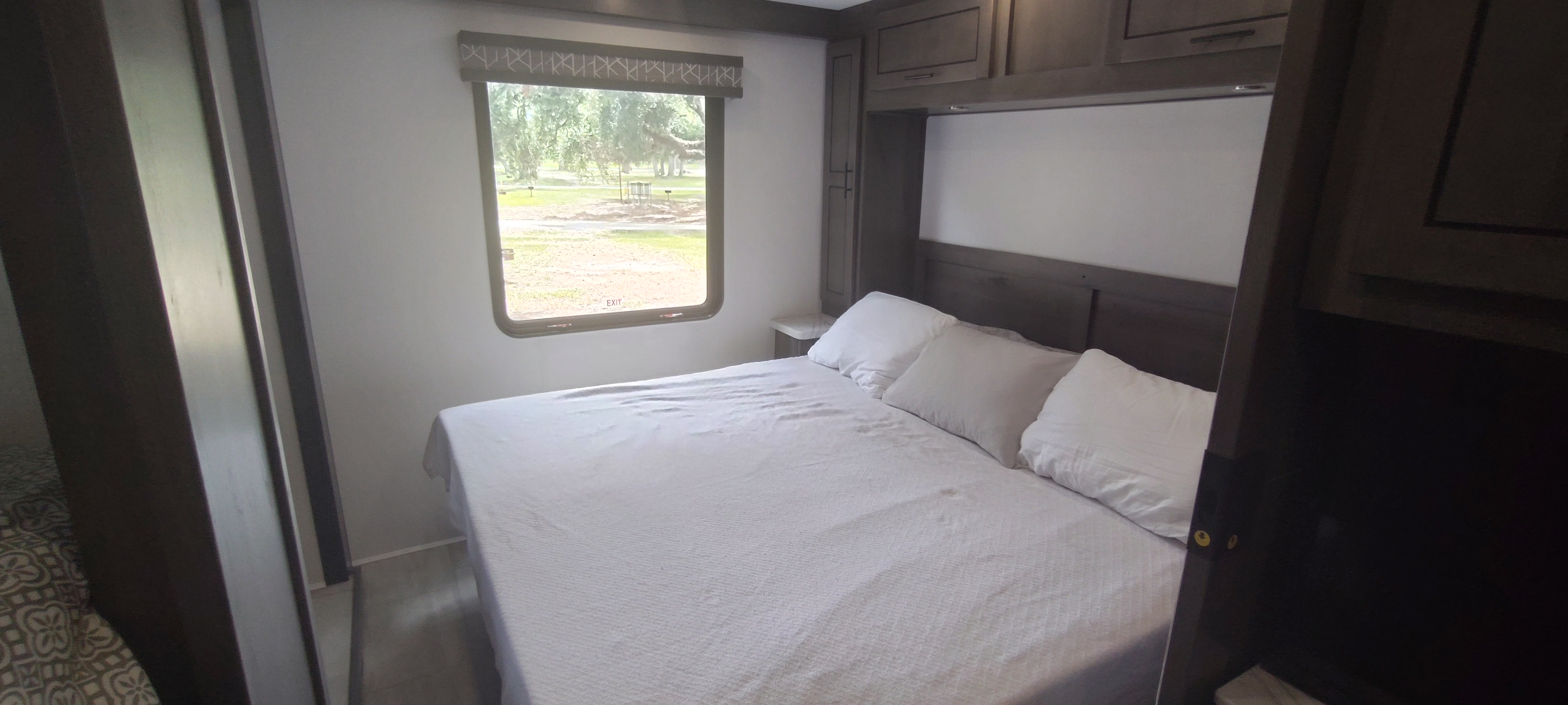 RV rental bedroom with a white-linened bed, overhead cabinets, and a window view of an outdoor park setting.. FR3 FR3 Motorhome 2024