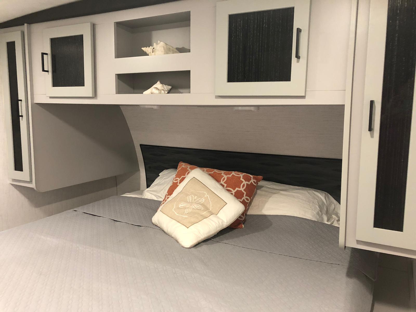 Cozy RV rental bedroom featuring a neat bed, decorative pillows, and white overhead cabinets with seashell decor.. Heartland RVs North Trail 2022