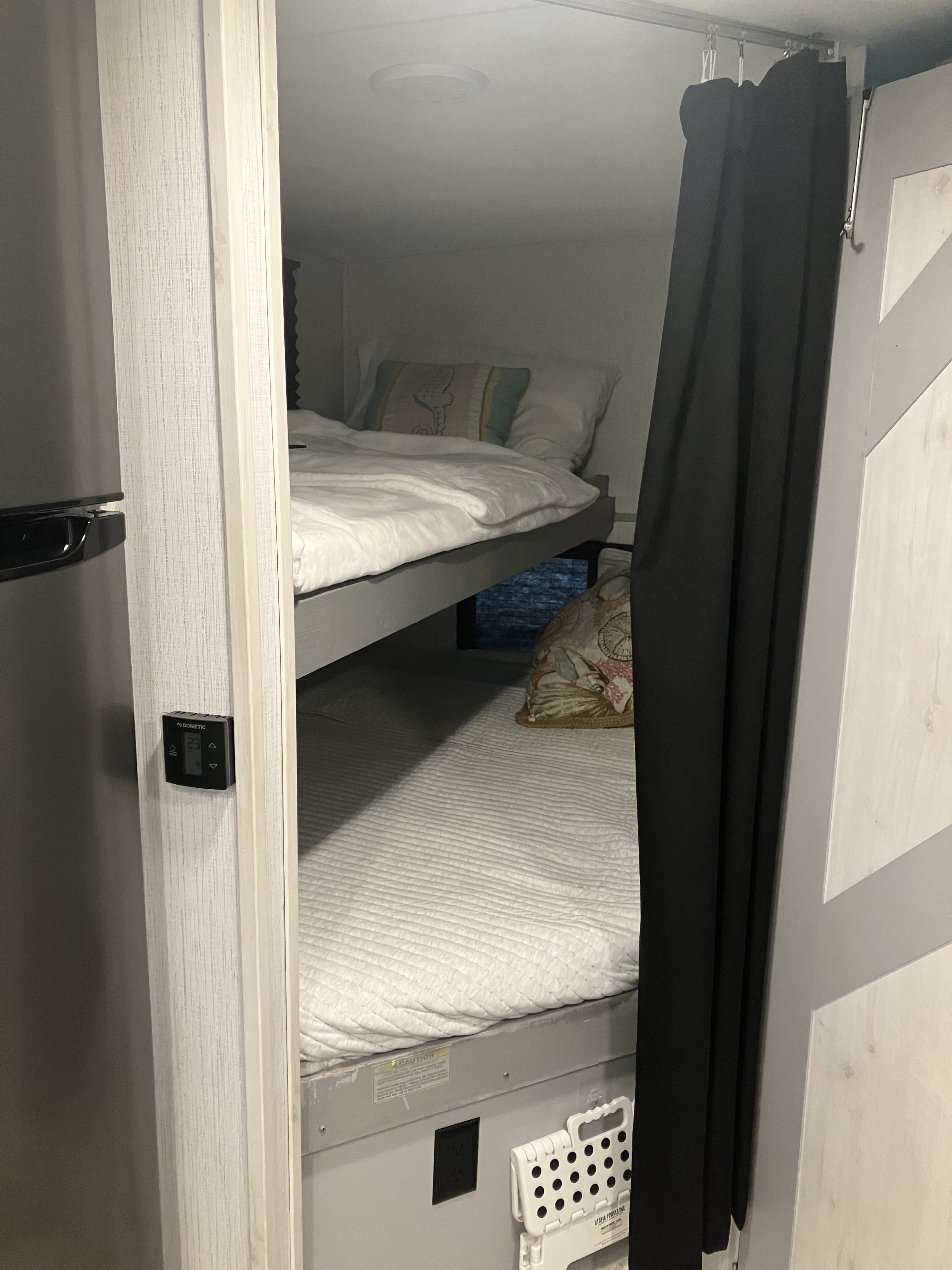 A view inside an RV rental showing a set of bunk beds with white bedding, a black privacy curtain, and a wall-mounted Dometic thermostat.. Heartland RVs North Trail 2022