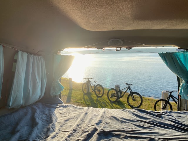 Serene RV rental view: cozy interior bed, ocean, and mountain bikes on grass. Ideal van life adventure.. Ford E350 Box Van 2003