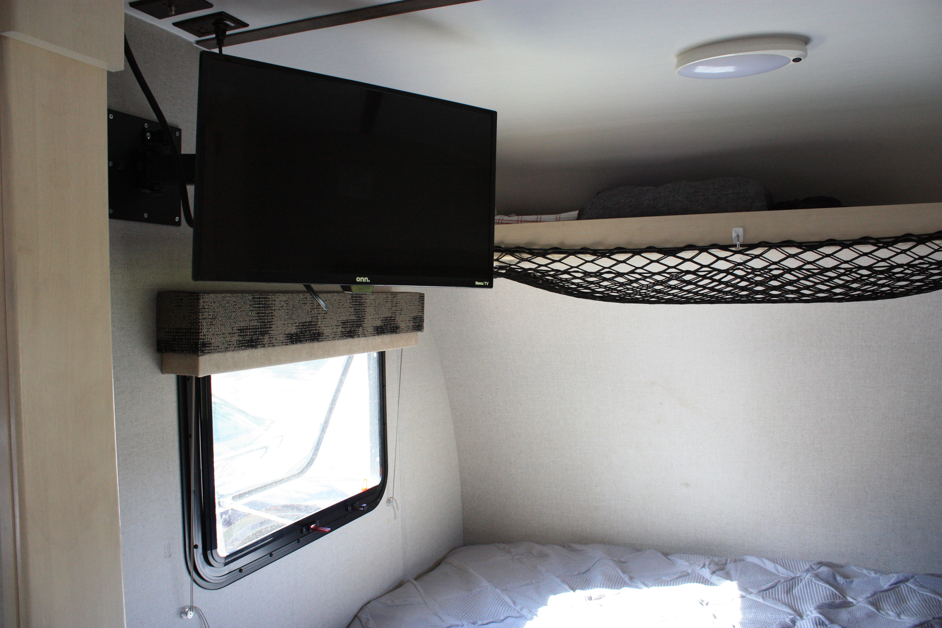 Inside a compact RV rental, showing a wall-mounted TV, window with valance, bed, and overhead net storage.. Winnie Drop Winnie Drop Trailer 2018
