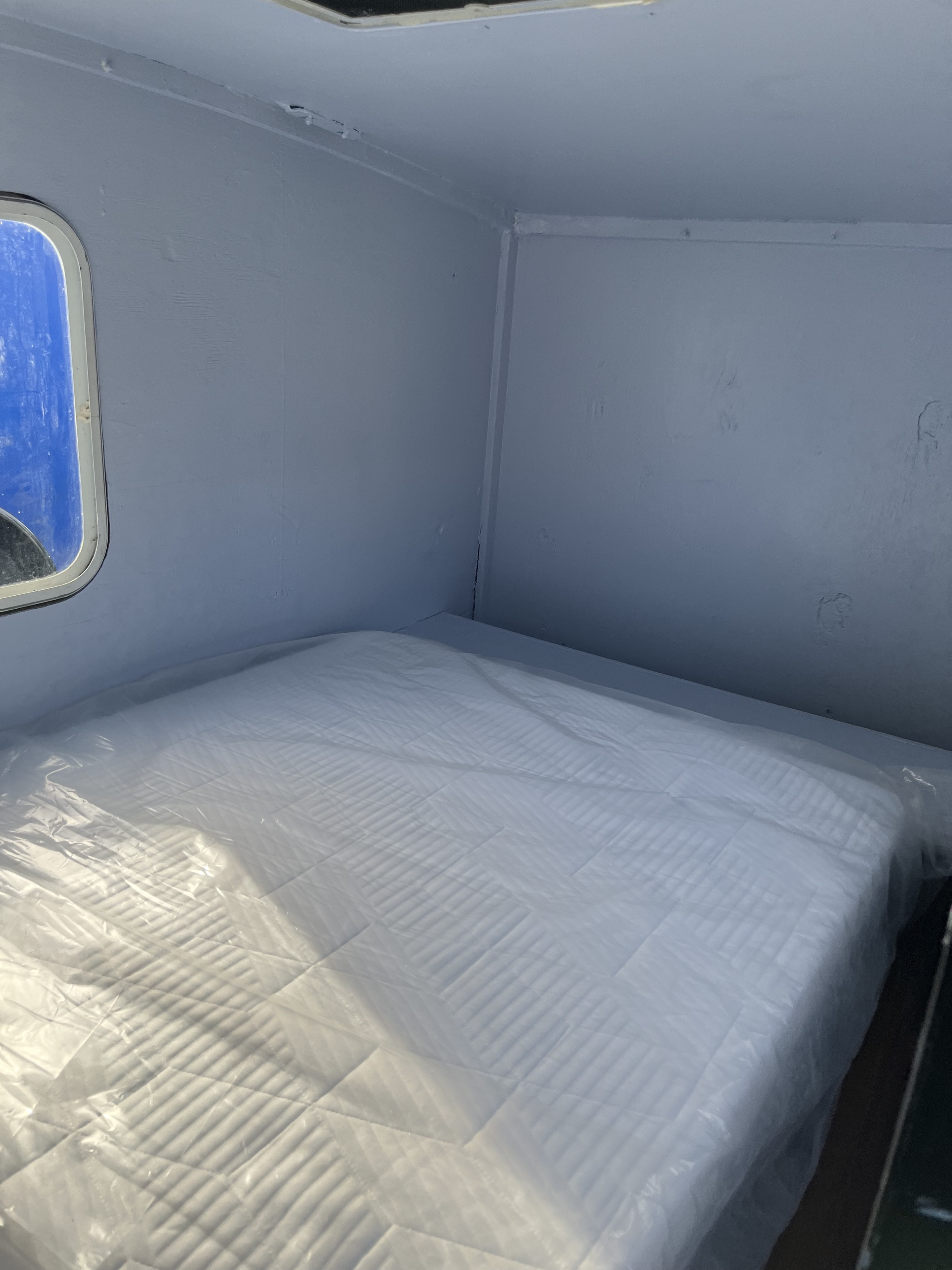 Inside an RV rental: a pristine mattress covered in clear plastic, set within a compact, light grey sleeping area with a small window.. Dutchmen AERO 400 2012