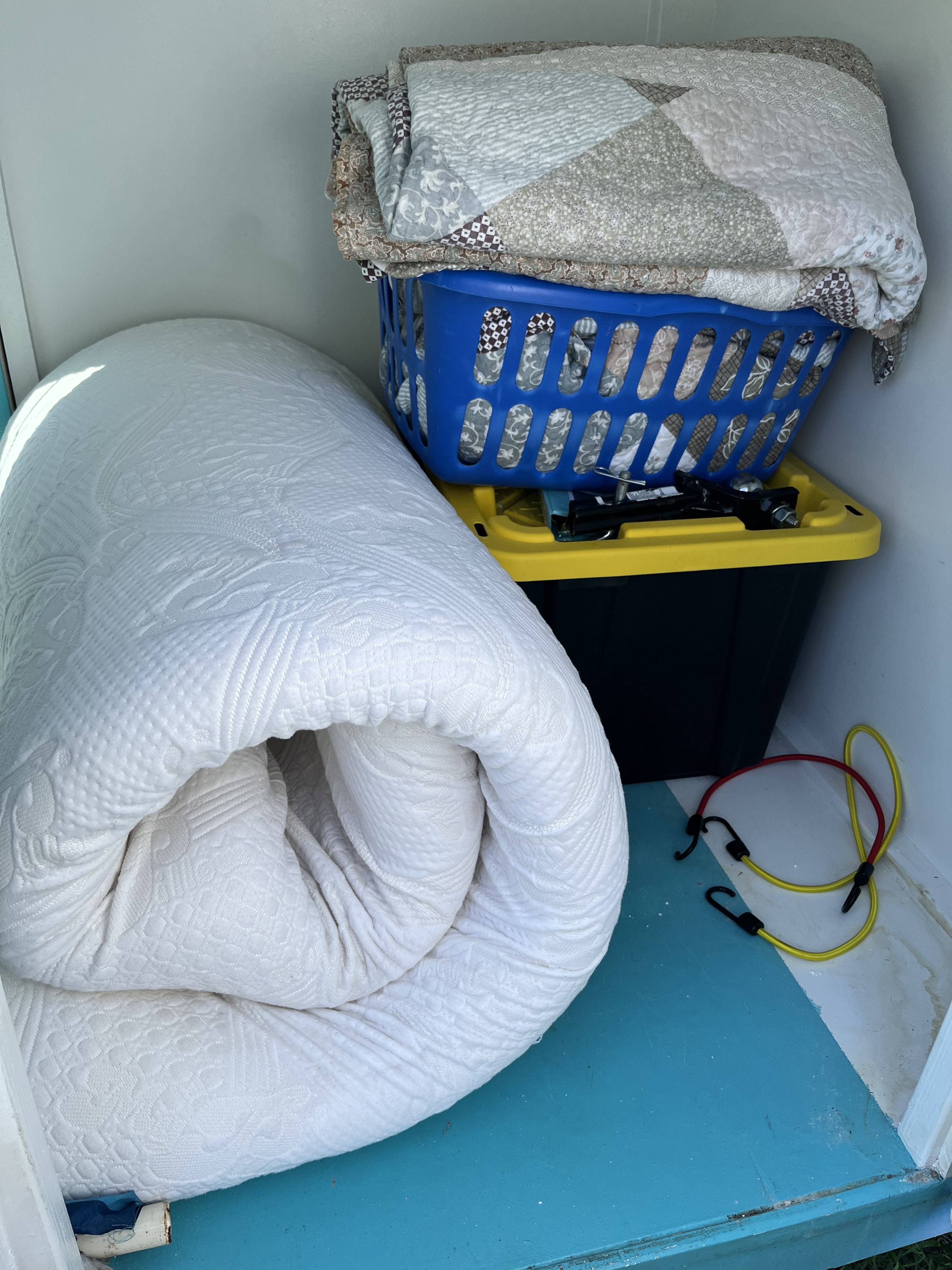 RV rental storage with rolled mattress, blankets in a basket, tools on a bin, and bungee cords on the floor.. YNGQ Box 2017