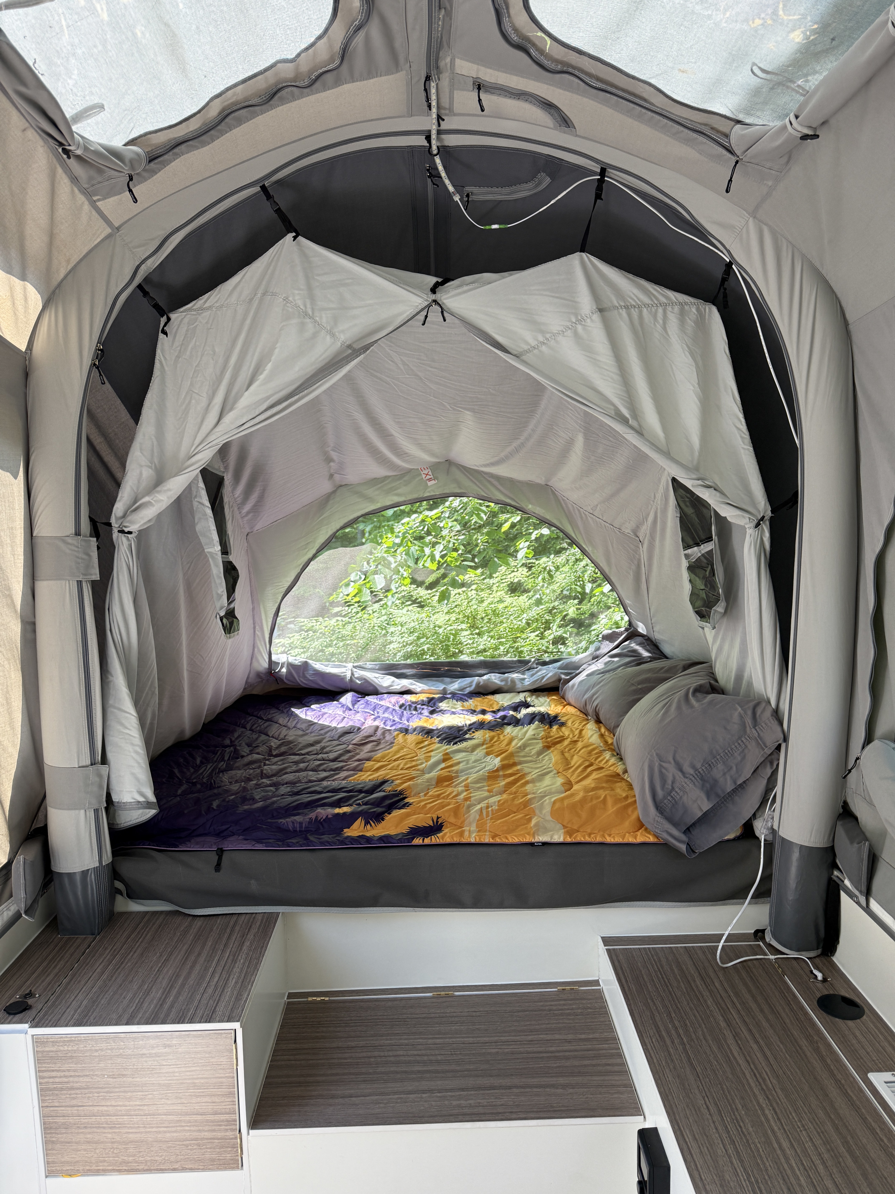 Inside an RV rental: Comfortable sleeping area with a vibrant comforter, nature views, and built-in storage solutions.. Opus Camper Opus Camper Popup Trailer 2023