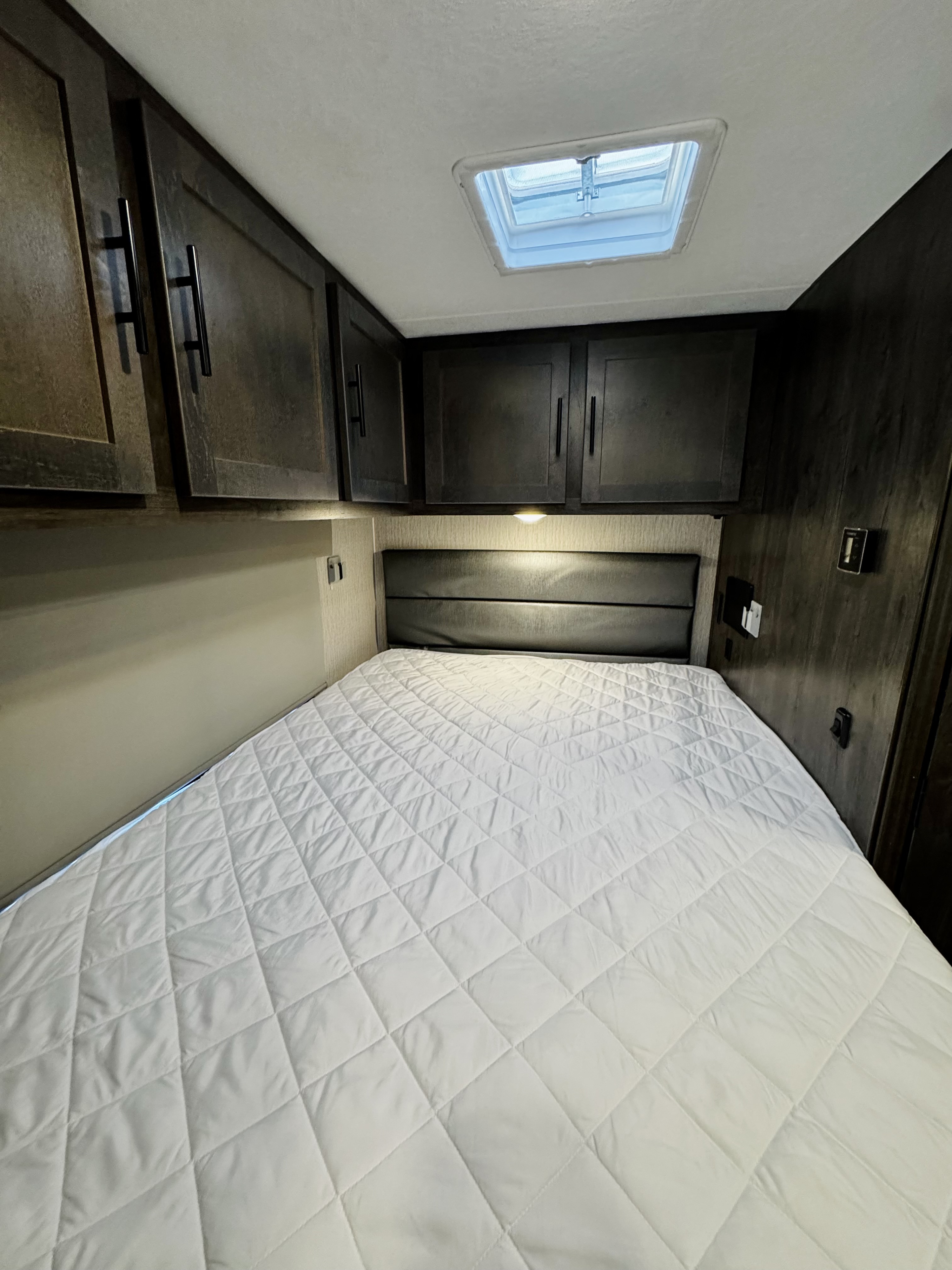 RV rental bedroom featuring a comfortable bed with a white quilted mattress pad, overhead cabinets, and a ceiling skylight.. Forest River Coachmen Prism LE 2023