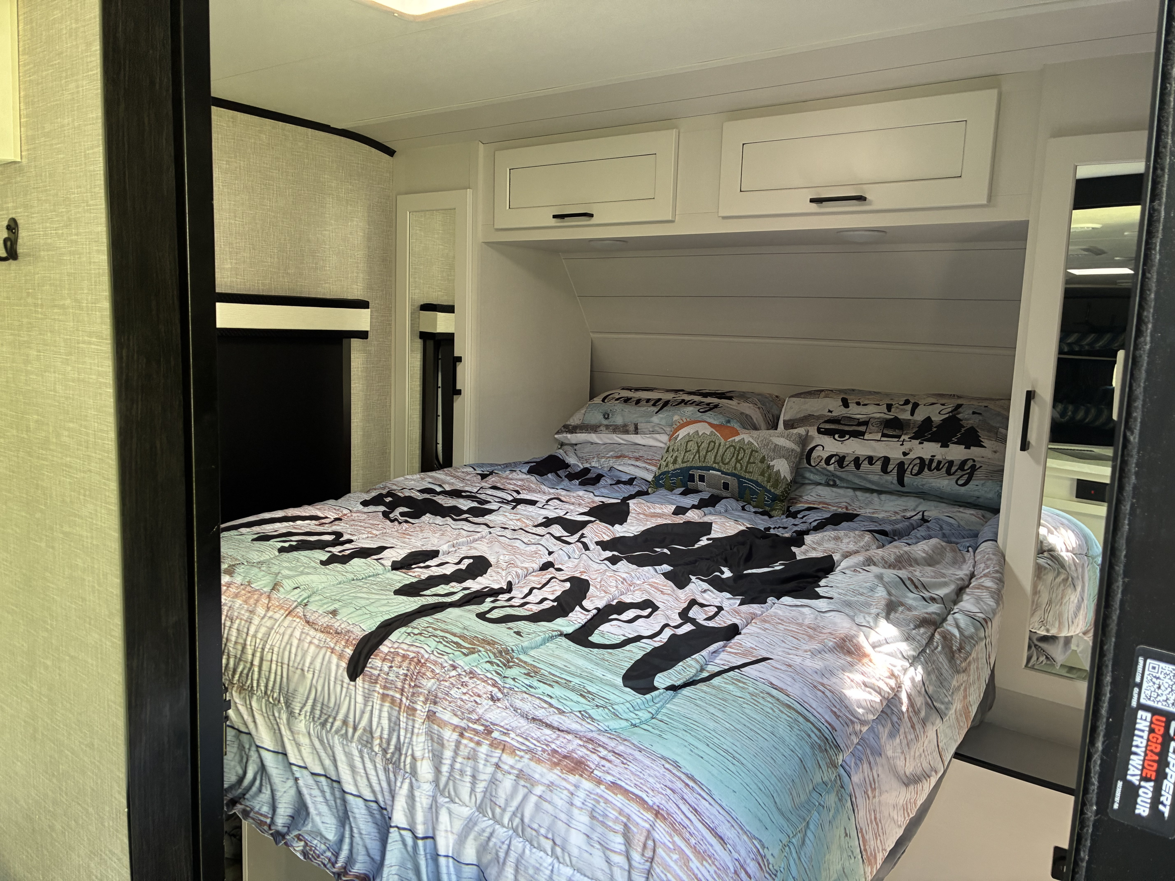 An RV rental bedroom with a rustic-themed bed, "Happy Camping" pillows, and built-in overhead cabinets for storage.. Jayco Jay Feather 2024