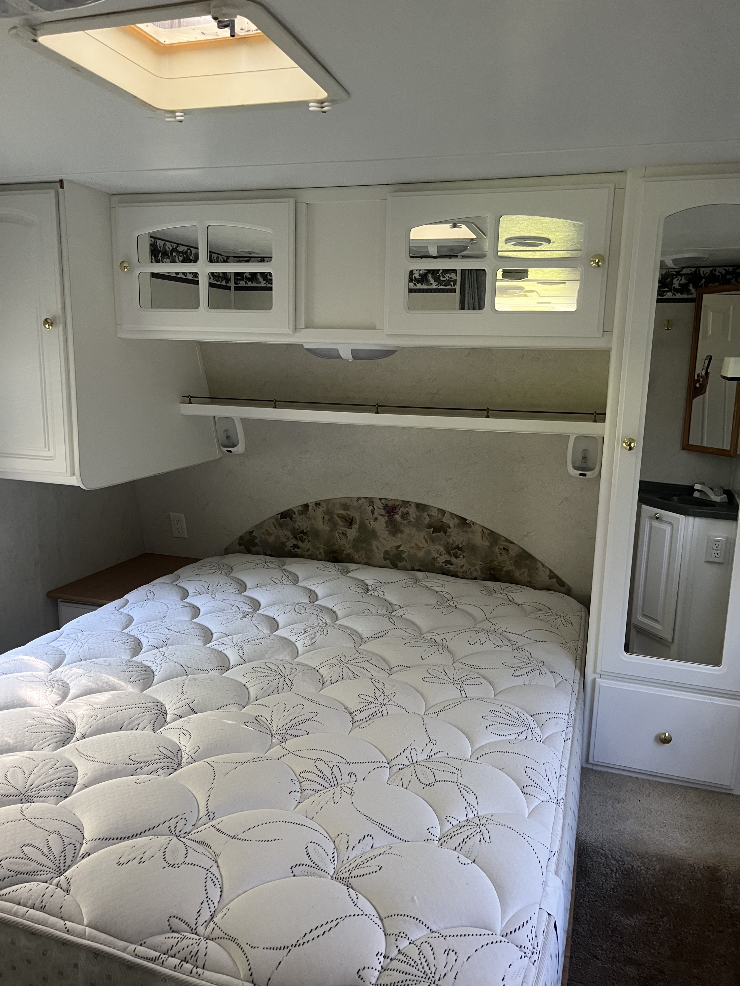 RV rental interior showcasing a bed with patterned mattress, overhead cabinets, and a skylight. Vanity area reflected.. Keystone RV LIteway Outback 2003