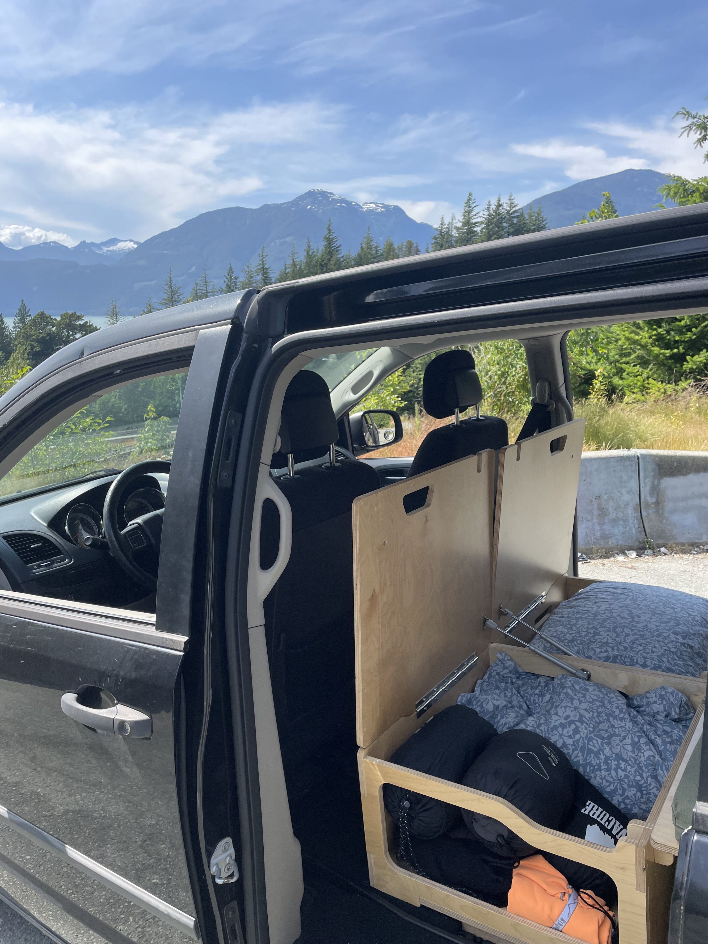 Black minivan camper conversion with custom wooden bed/storage inside, parked amidst mountains. A simple, practical RV rental option.. Dodge Grand Caravan 2014