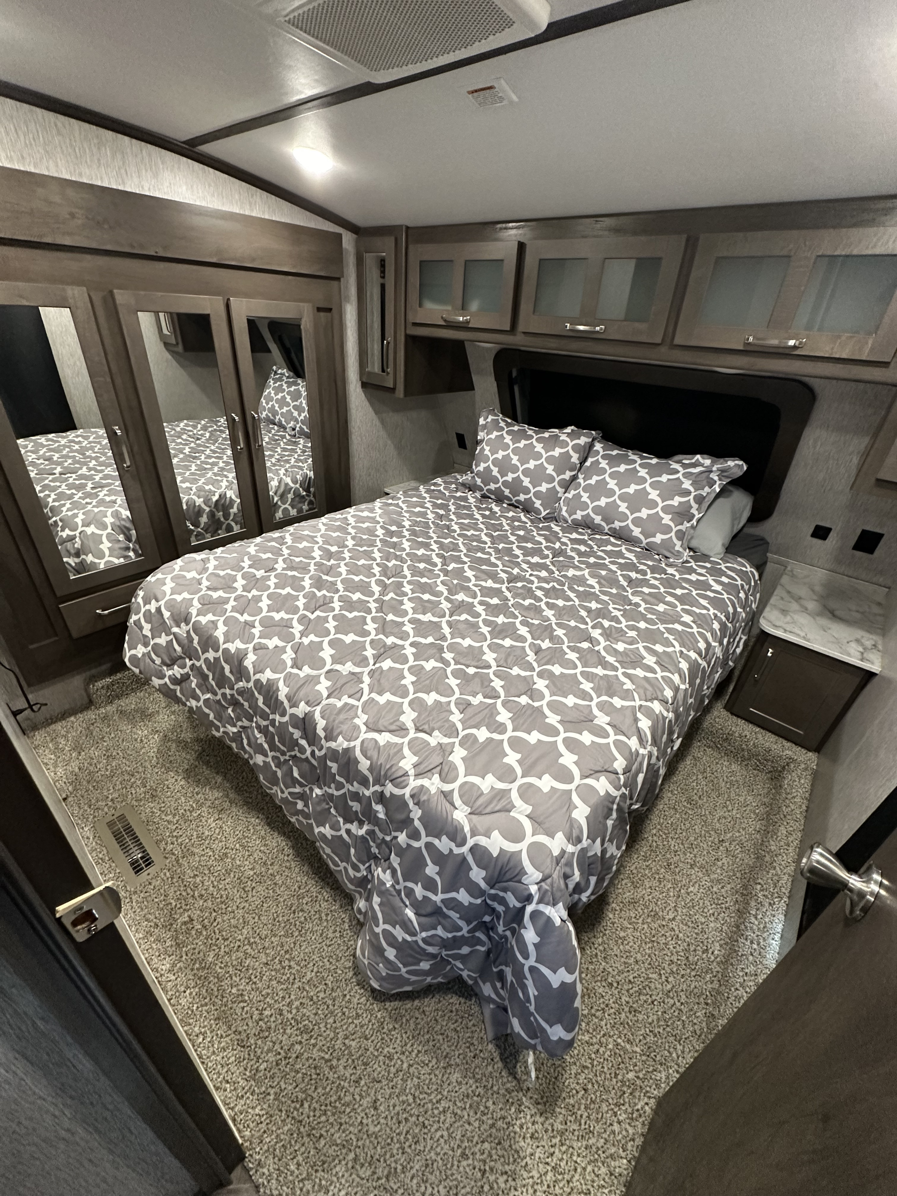 Inside a comfy RV rental bedroom: patterned bed, mirrored closet, overhead storage, and warm lighting details.. Astoria 1500 2021