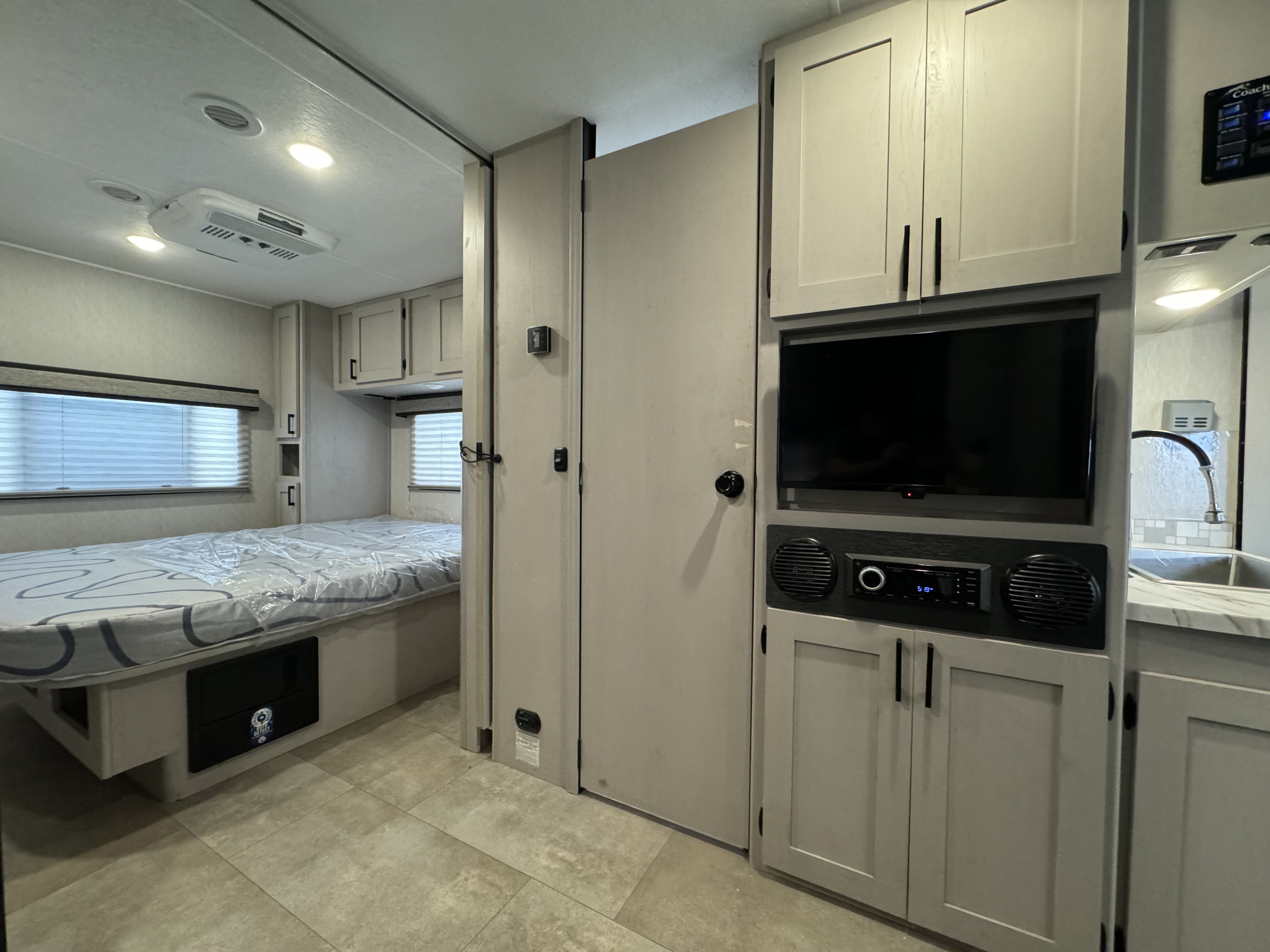 Clean RV rental interior featuring a bright bedroom with bed, window, and storage, connecting to a living space with TV and sink.. Coachmen 30BH 2023