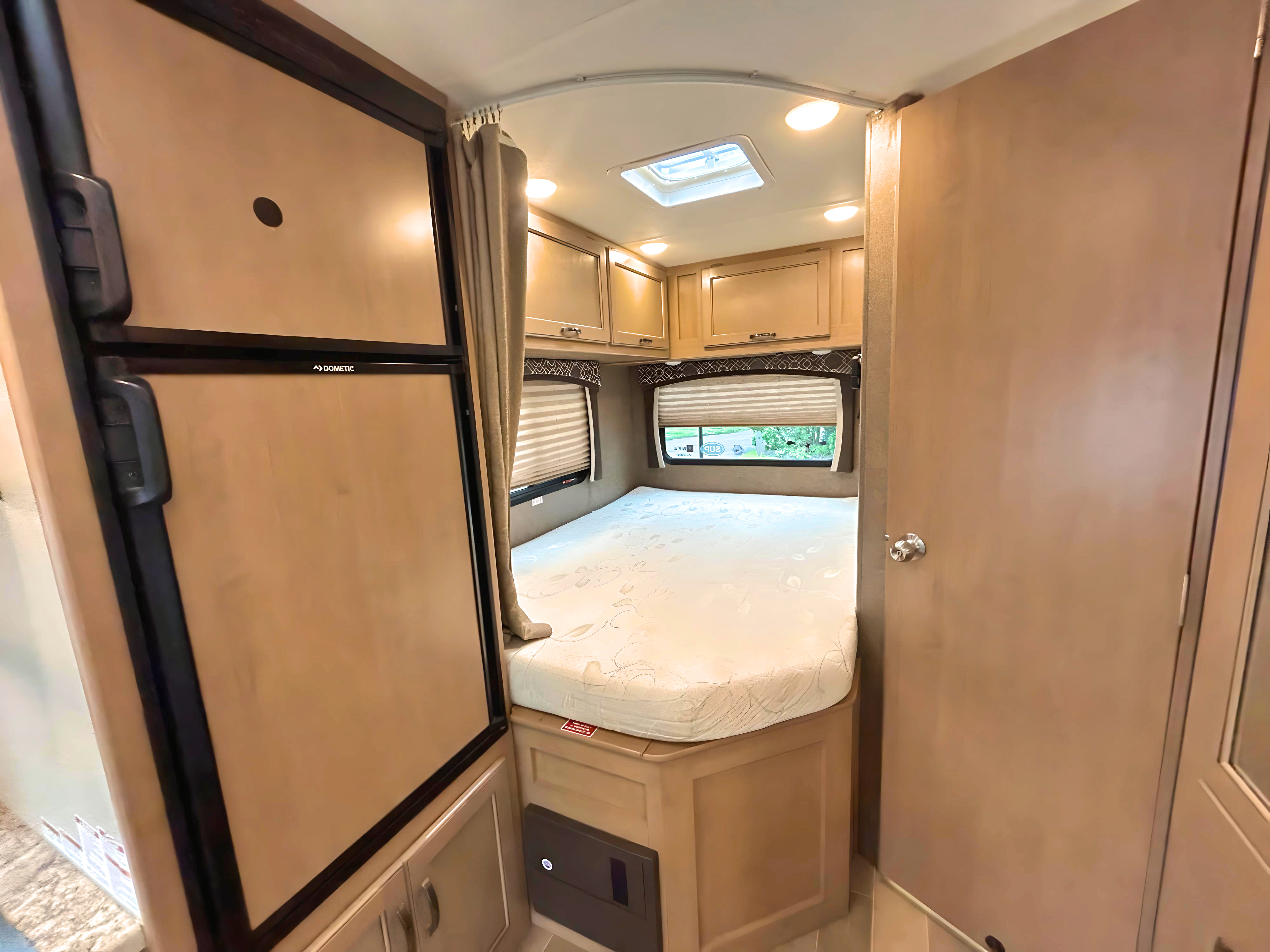 This RV rental interior shows the bedroom with a bed, windows, and overhead cabinets, beside a Dometic refrigerator.. Thor Chateau 2019