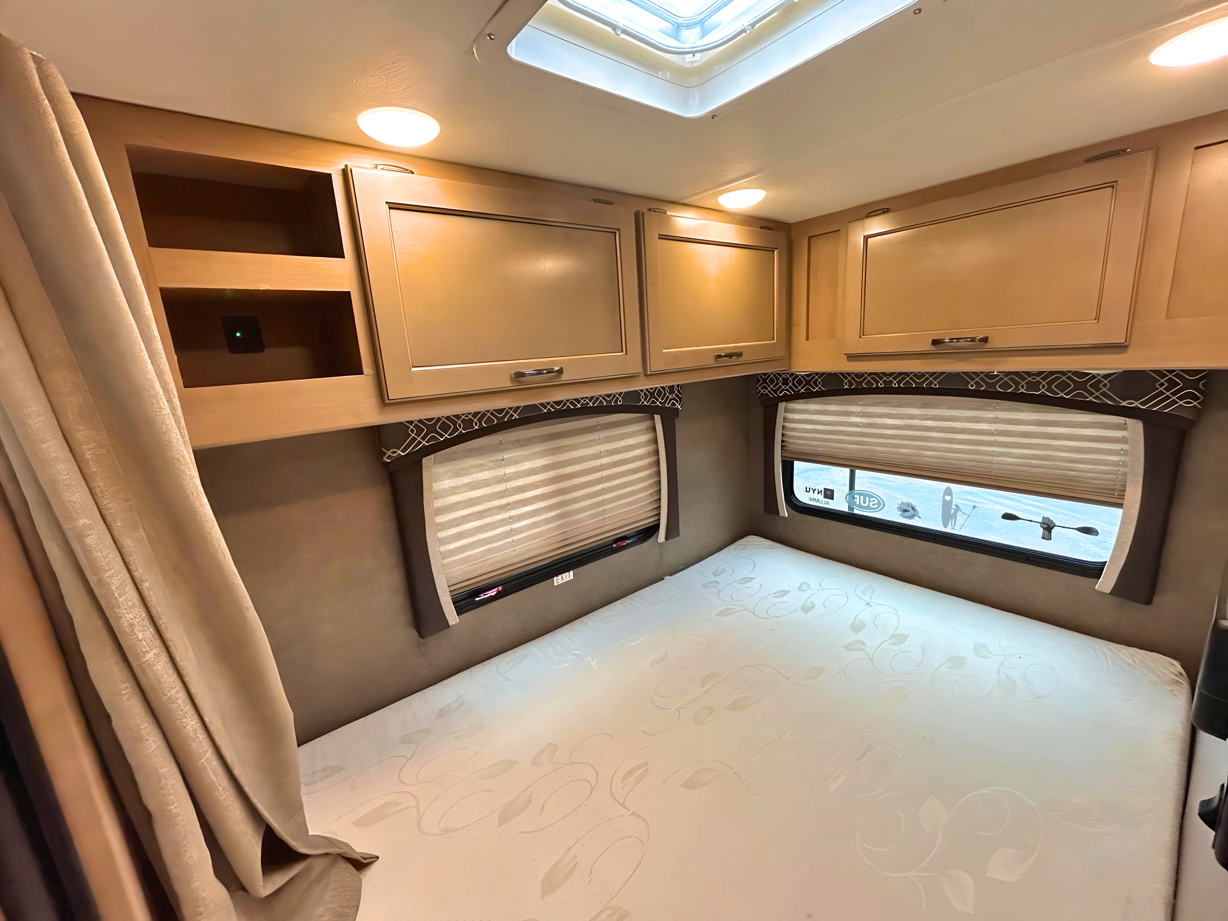 Inside an RV rental: a bright bedroom with a patterned mattress, overhead cabinets, windows with blinds, and a ceiling skylight.. Thor Chateau 2019