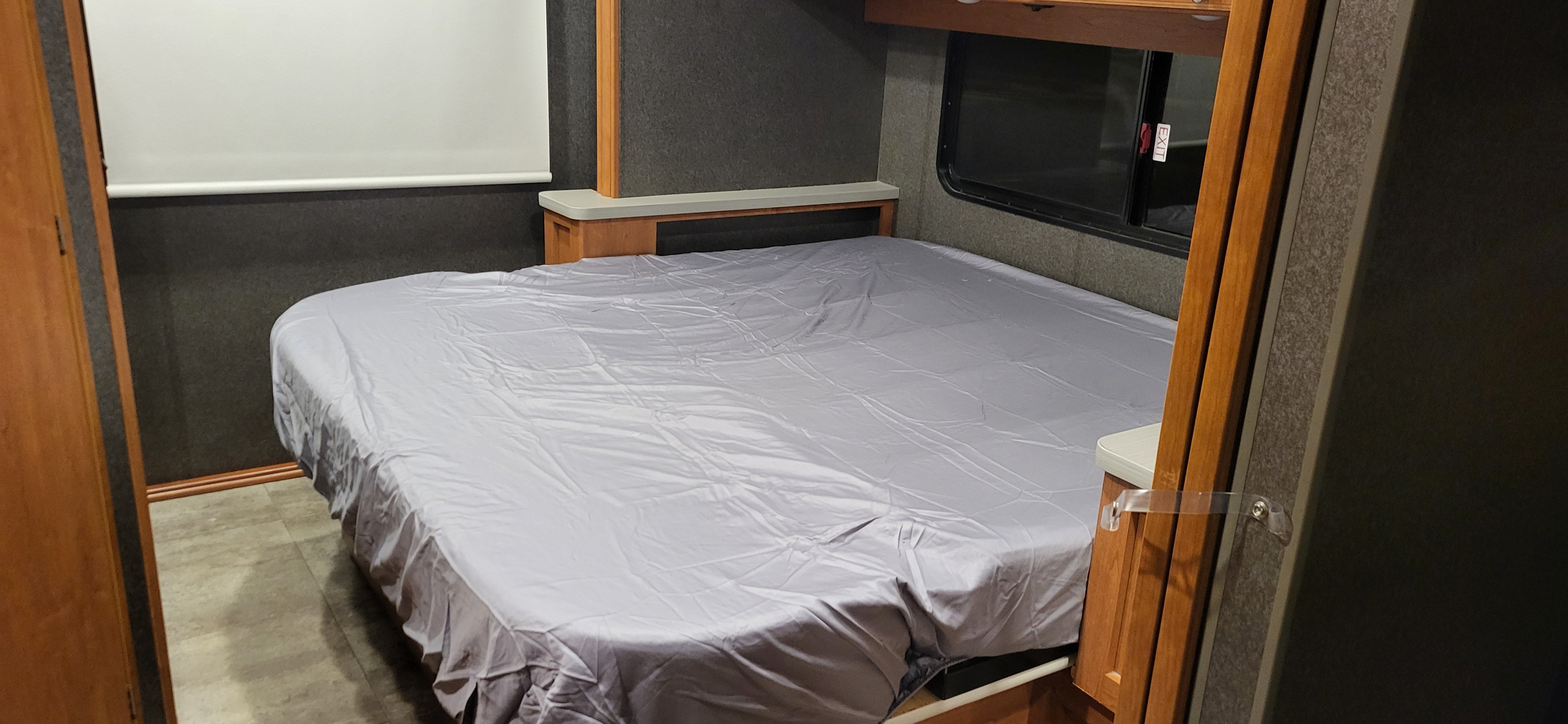 RV rental bedroom features a bed with grey cover, a side window, and built-in headboard storage.. Winnebago Vista 2018