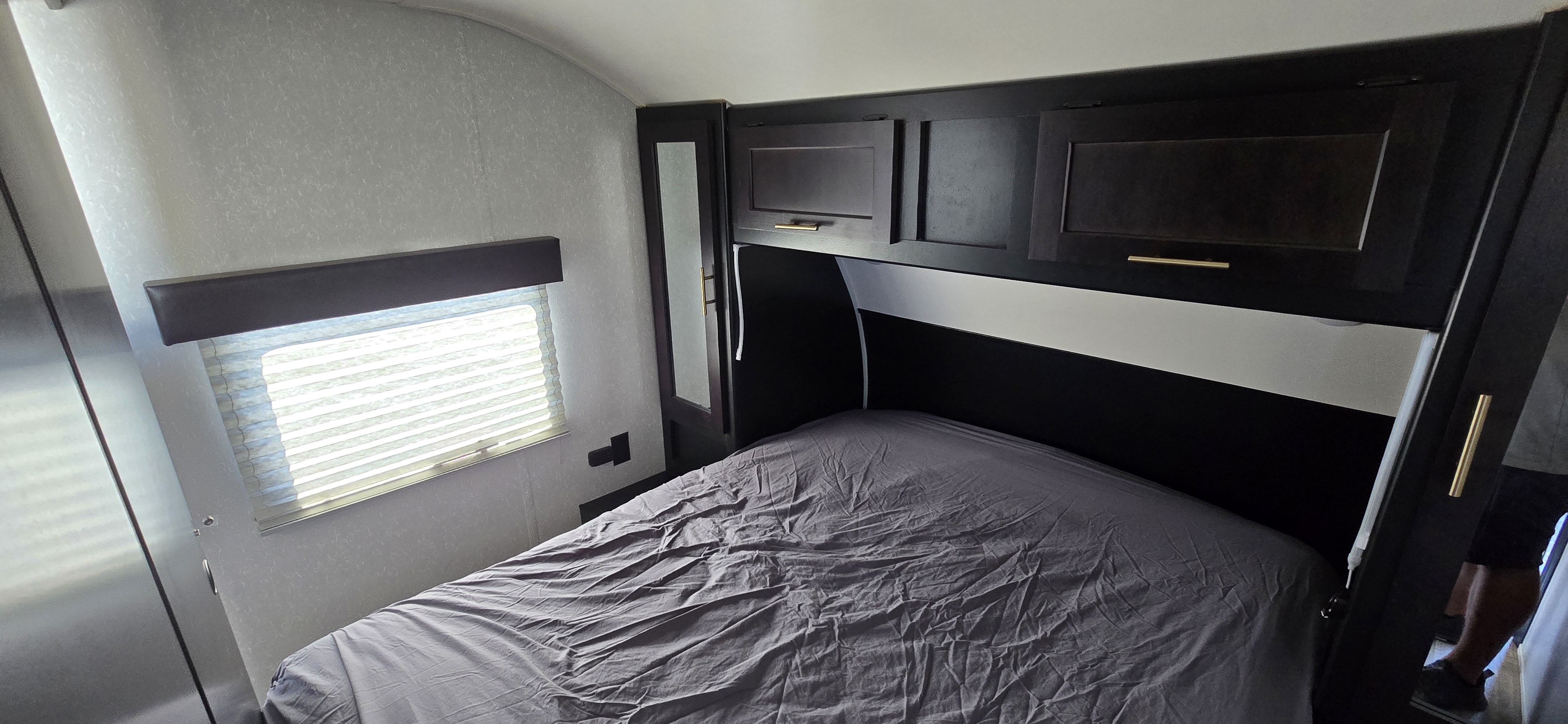 Cozy RV rental bedroom featuring a grey bed, overhead cabinets, and a window with blinds.. Forest River Cherokee Grey Wolf 2021