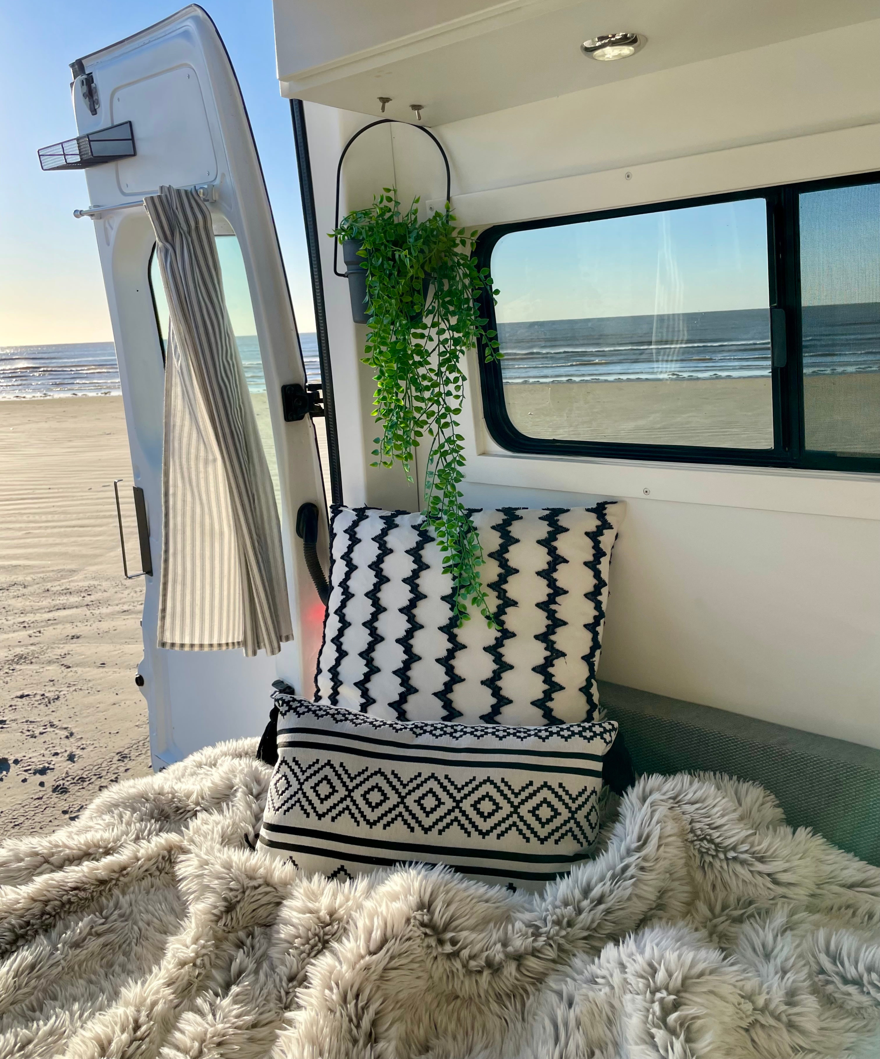 Cozy RV rental interior with black & white pillows & furry blanket, offering stunning ocean & beach views from the open back.. Mercedes-Benz Sprinter 2016