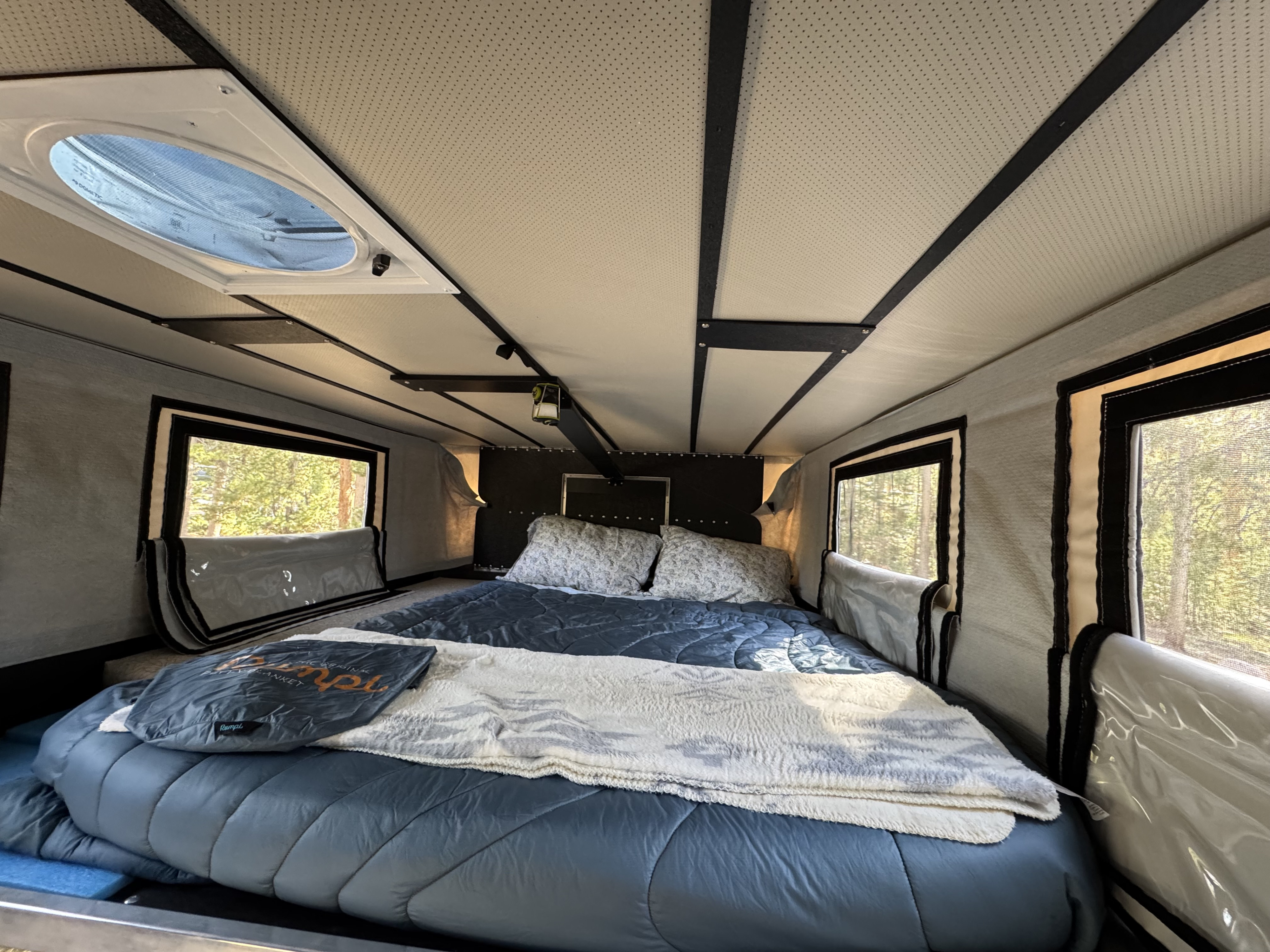 Inside a comfortable RV rental bedroom, featuring a large bed, side windows with tree views, and a Dometic ceiling vent.. Four Wheel Campers Shell Models 2020