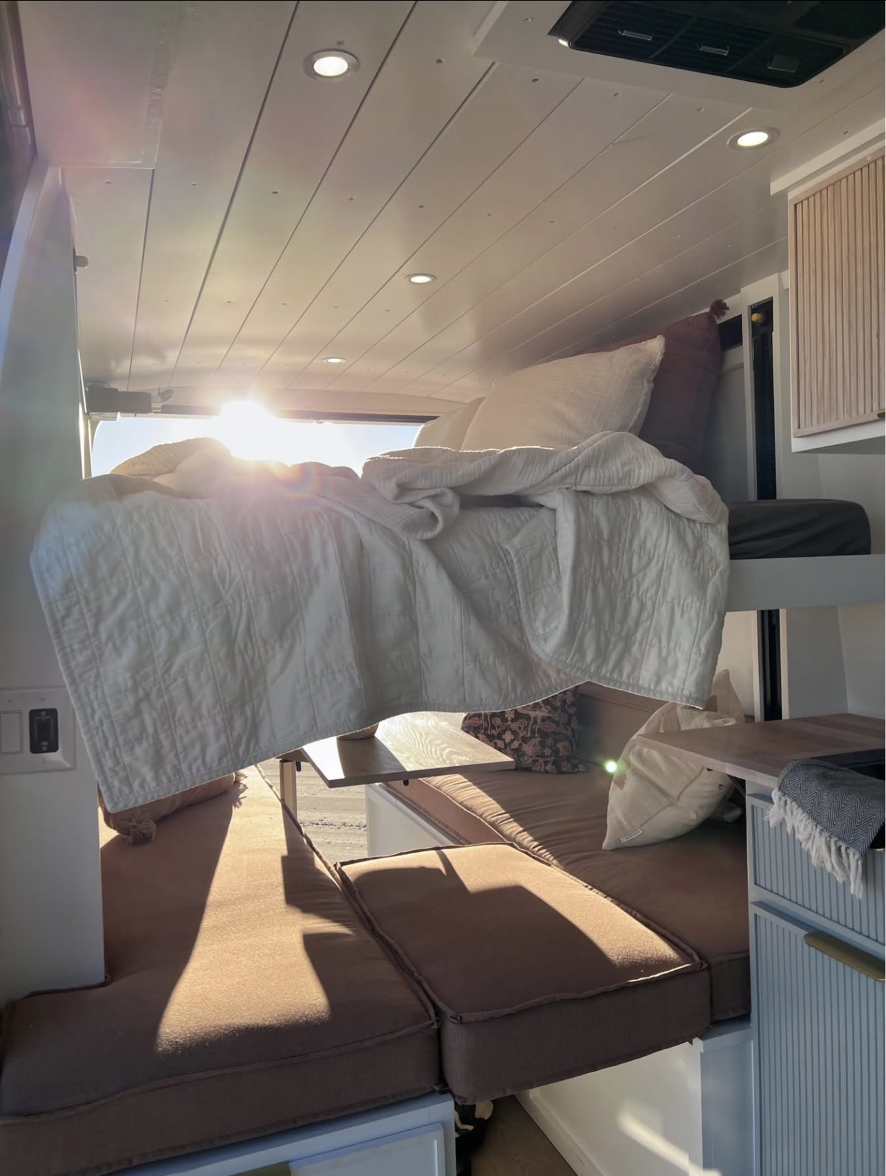 This bright RV rental interior showcases a suspended bed with white bedding floating above brown cushioned seating, bathed in sunlight from the rear.. Mercedes-Benz Sprinter 2021