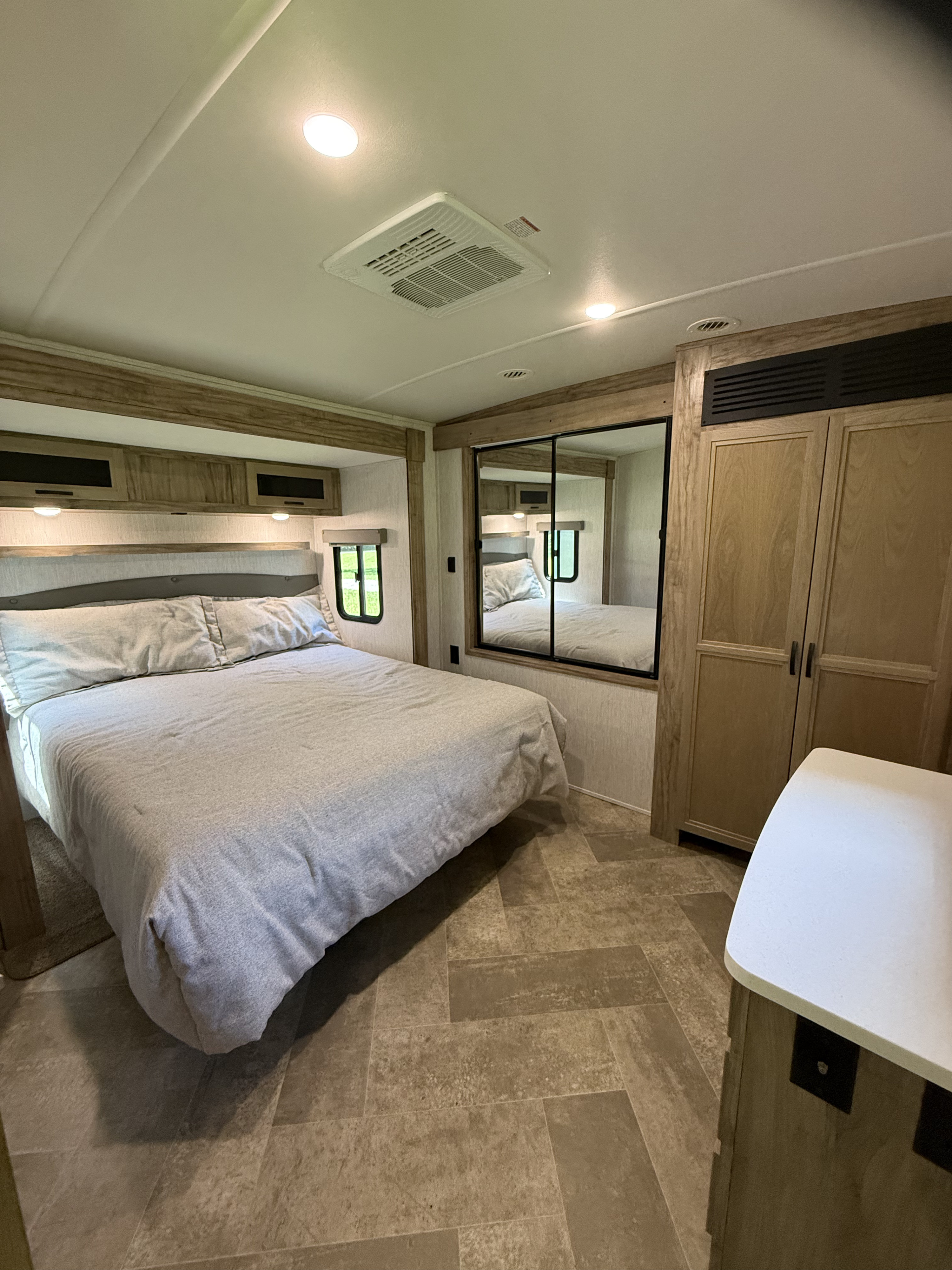Inside a modern RV rental bedroom: bed with gray linens, overhead cabinets, mirror, closet, and AC unit.. Palomino SolAire 320 TSBH 2022